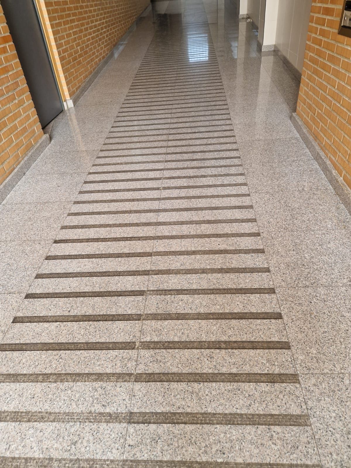 Pasillo con suelo moteado; el centro tiene bandas horizontales oscuras, flanqueado por paredes de ladrillo y una puerta.
