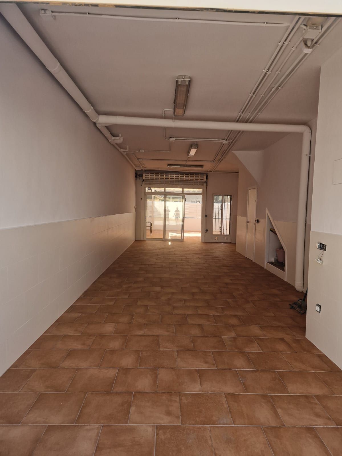 Espacio comercial vacío con piso de baldosas marrones, paredes blancas y puertas de vidrio en el extremo más alejado.