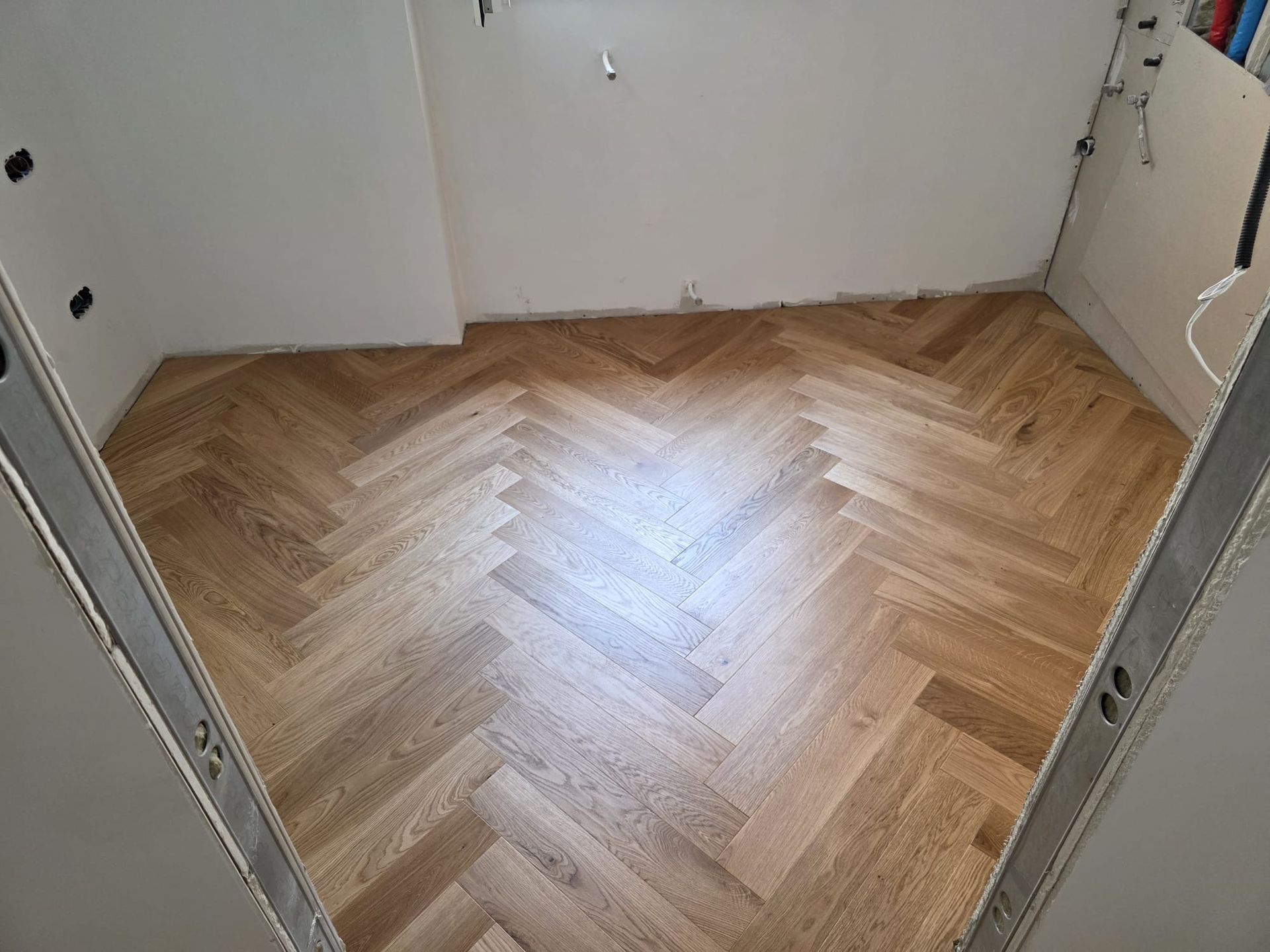 Piso de madera en espiga en una habitación, con paredes blancas y marcos de puertas sin terminar.
