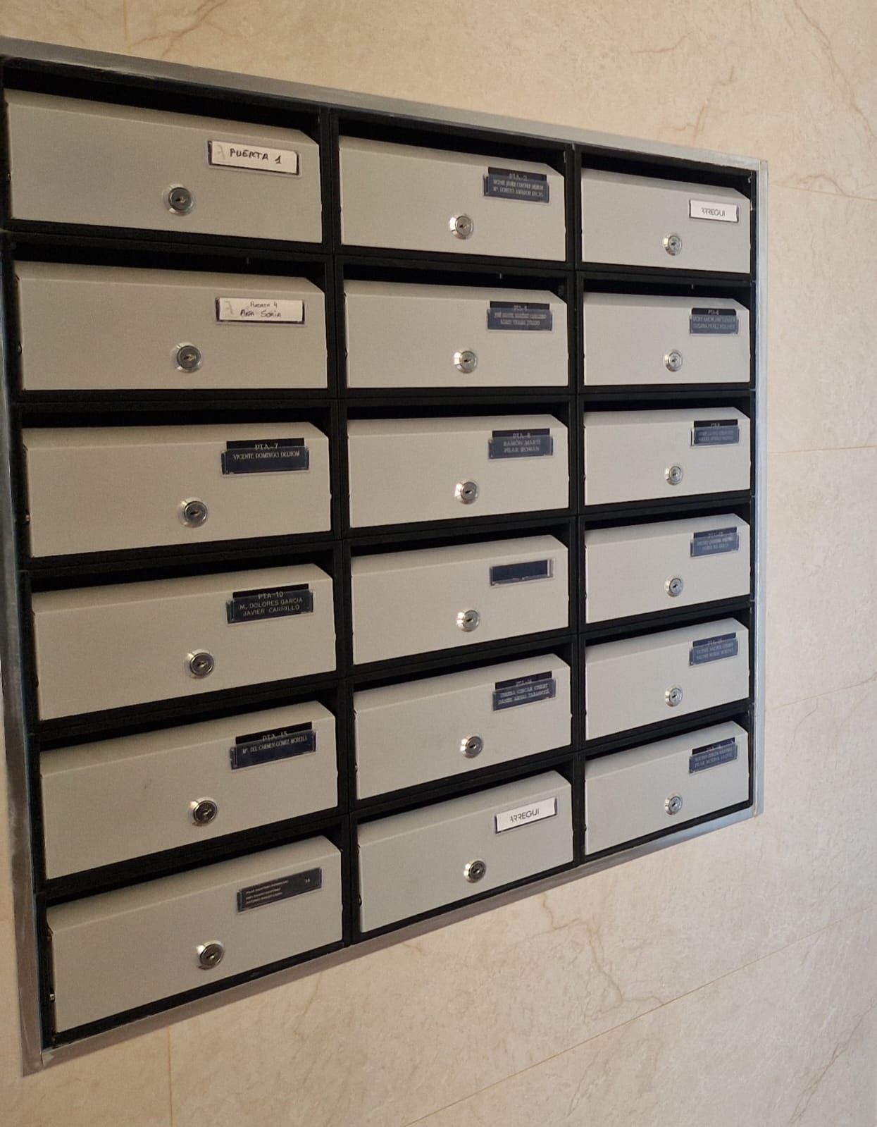 Un banco de buzones grises montados en la pared, cada uno con cerradura y placa con nombre, en un edificio.