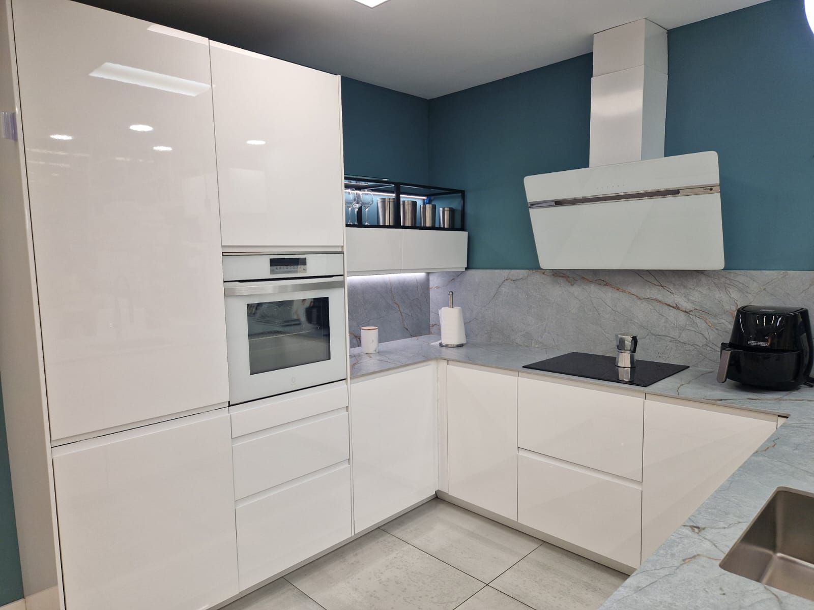 Cocina blanca moderna con horno empotrado, anafe y campana extractora contra pared verde azulado.