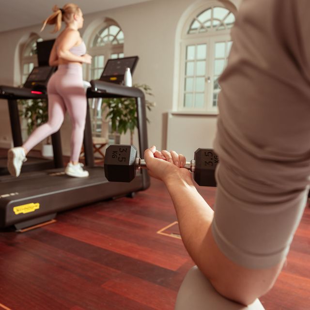 Eine Person hebt im Fitnessstudio eine Hantel; eine Frau läuft auf dem Laufband.