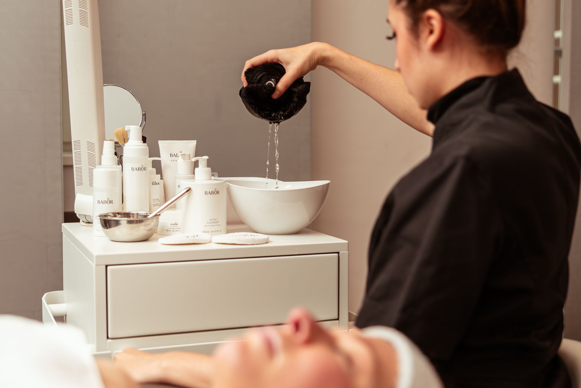 Facial treatment: A beautician is pouring water from a bowl. Skin care products are on a table next to her.