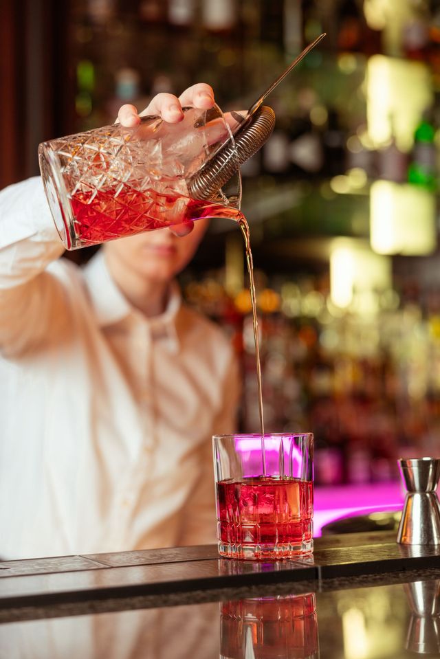 Ein Barkeeper schenkt an einer Bar einen roten Cocktail in ein Glas ein.