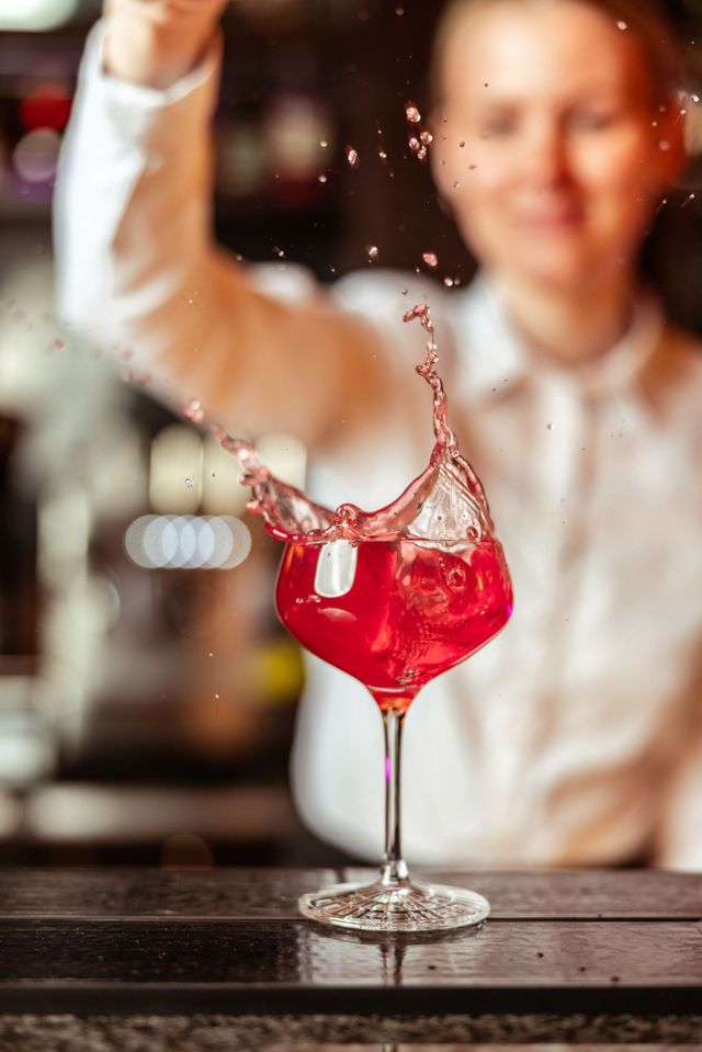Ein roter Cocktail spritzt in einem Stielglas auf einer Bar; dahinter steht ein verschwommener Barkeeper.