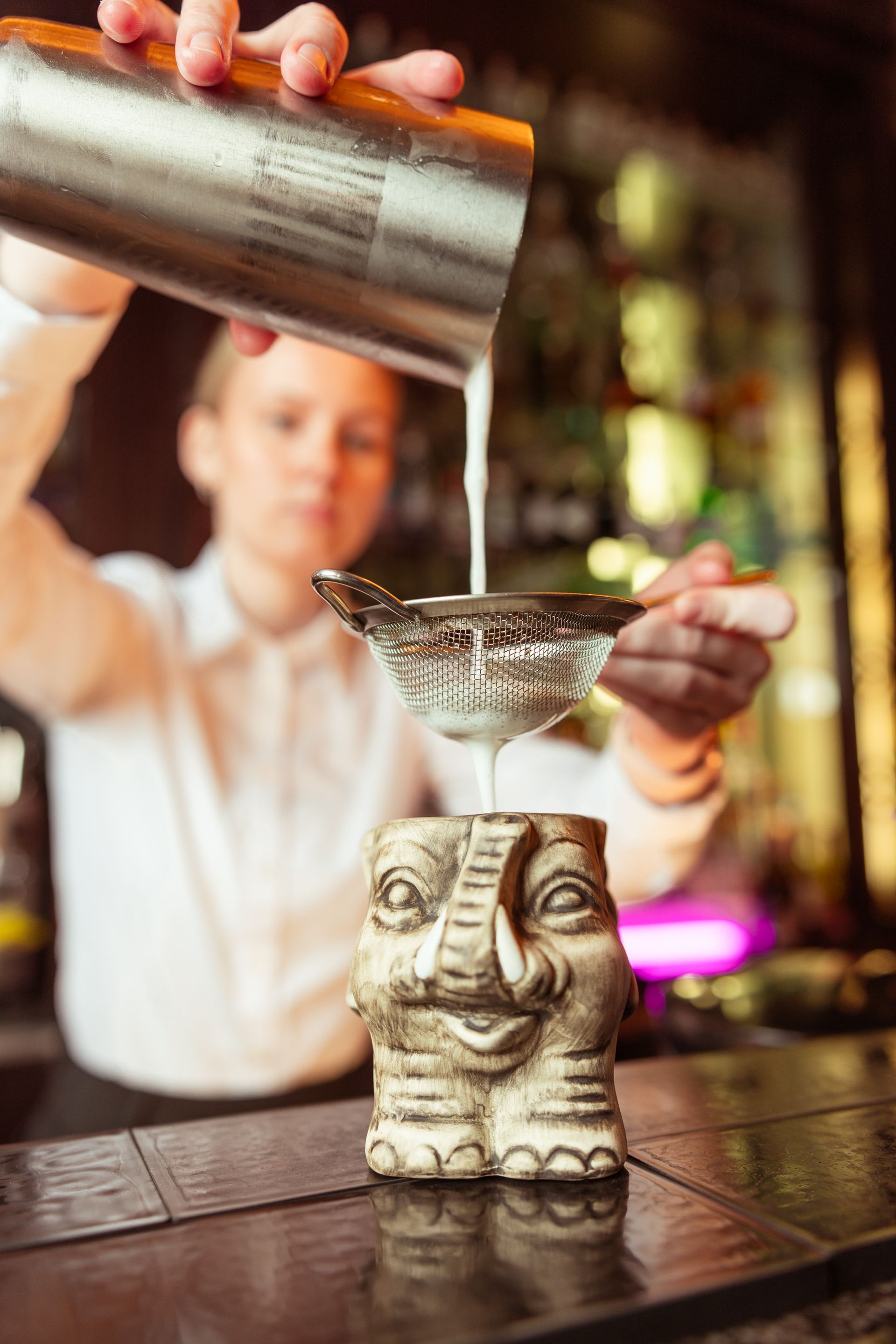 A bartender pours a cocktail from the shaker through a strainer into an elephant-shaped cup on the bar.