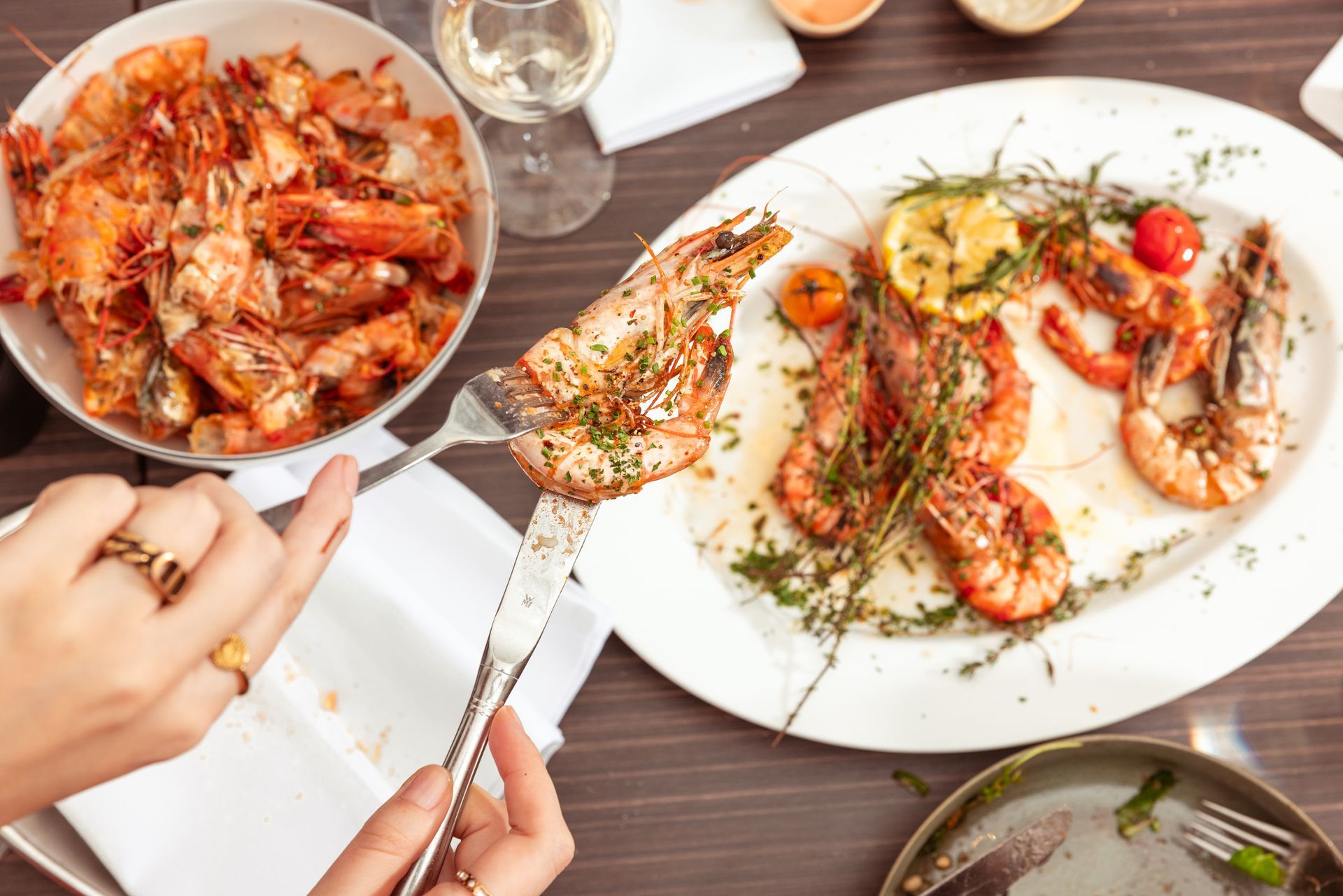 Eine Person hält mit einer Gabel gegrillte Garnelen auf einem Tisch mit Garnelengerichten, Wein und weiteren Zutaten.