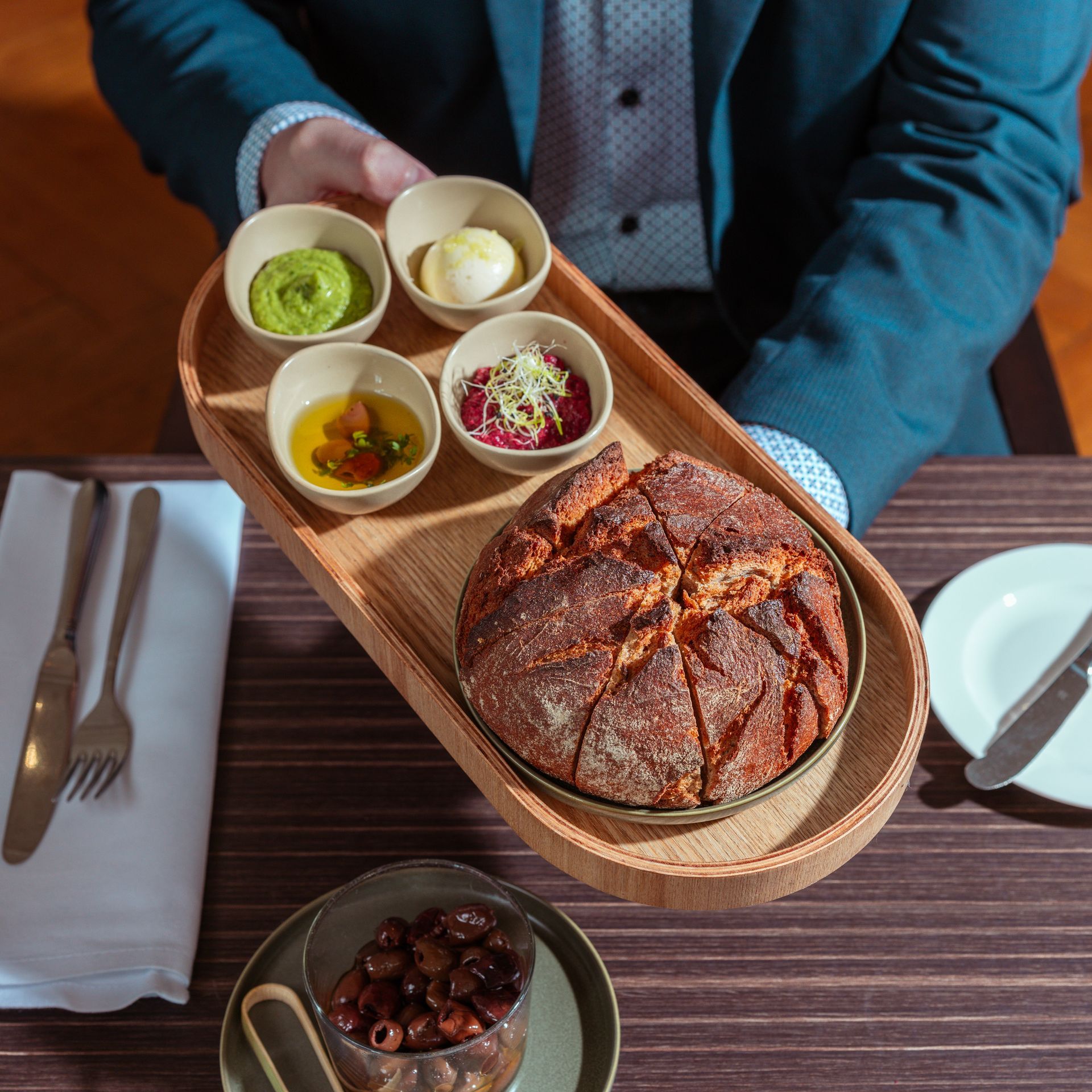 Eine Person hält ein Holztablett mit Brot und Dips; gedeckter Esstisch.