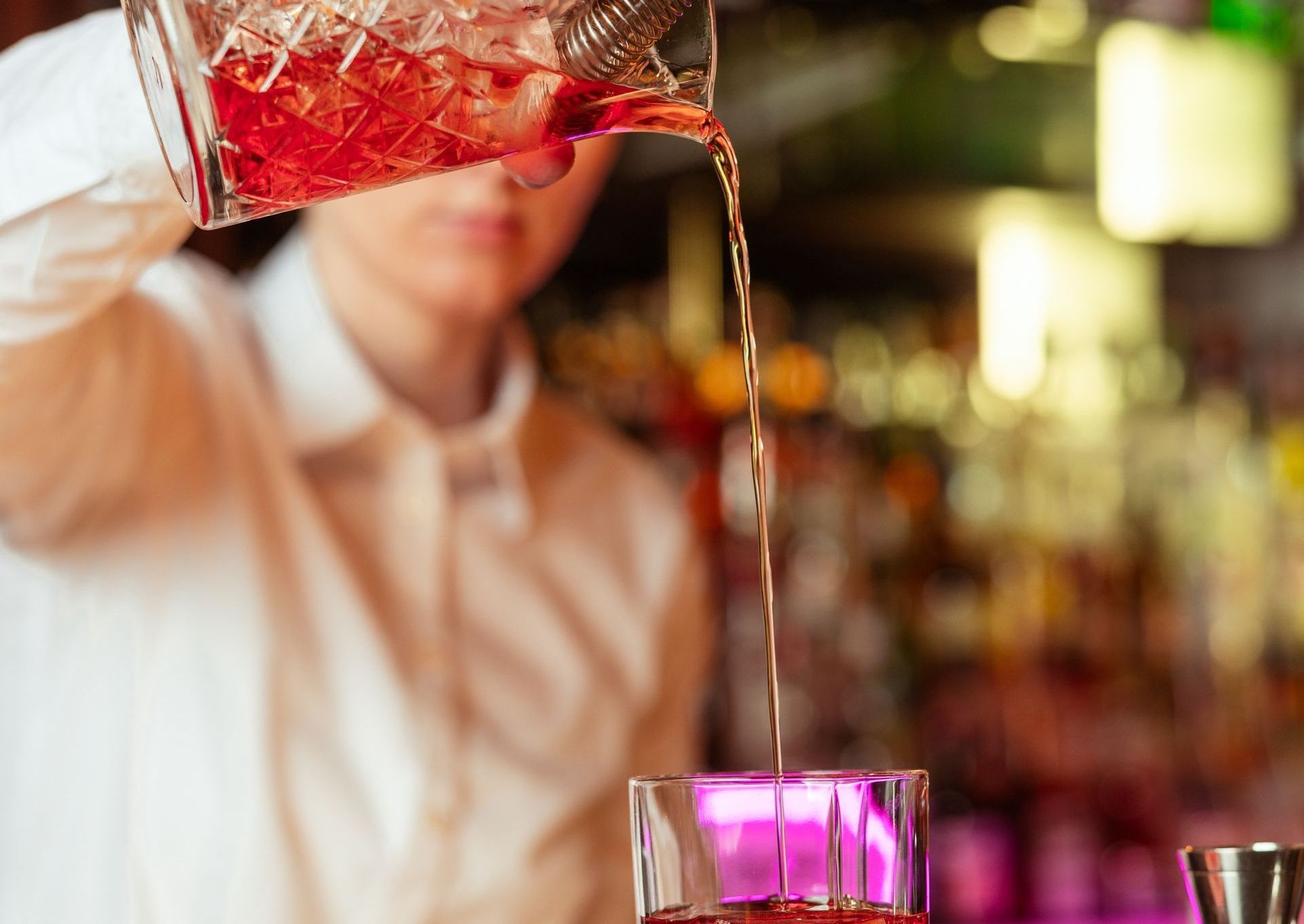 A bartender pours a red drink from a shaker into a glass at a bar.