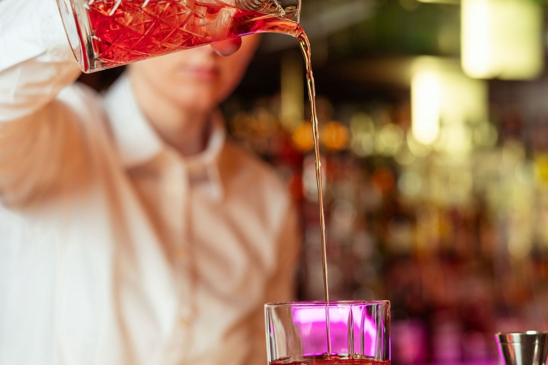 A bartender pours a red cocktail into a glass at a bar.