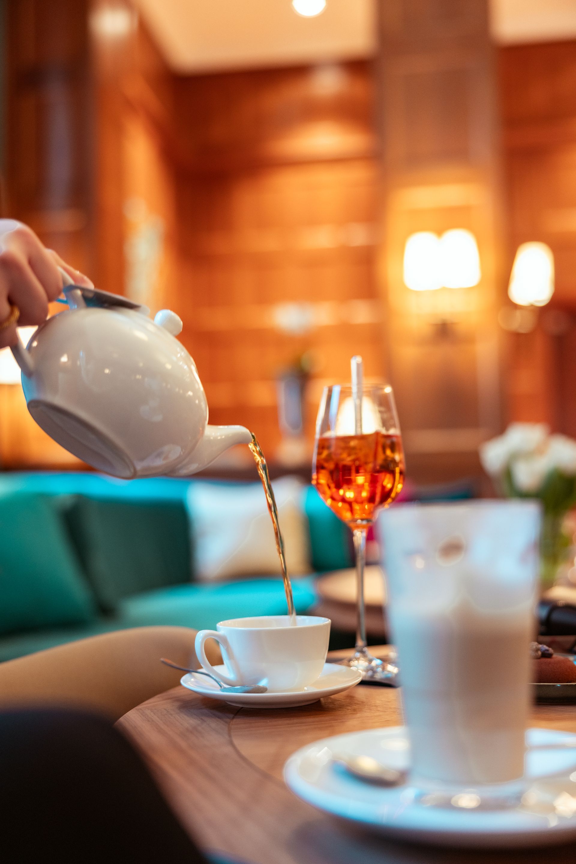 Jemand gießt Tee in eine weiße Teetasse an einem Tisch mit anderen Getränken in einem warmfarbigen Raum.