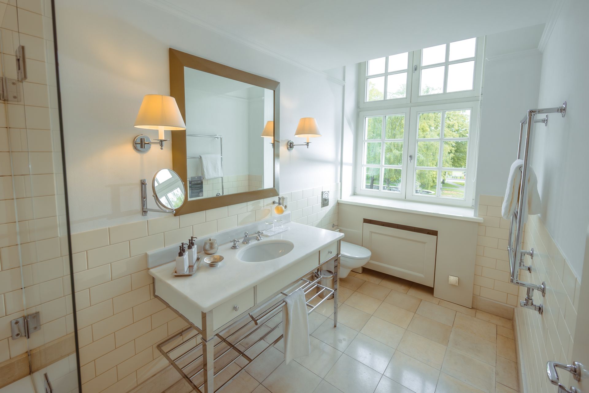 A bright, modern bathroom with cream-colored tiled walls, a vanity with a marble countertop, a large mirror, and a window.