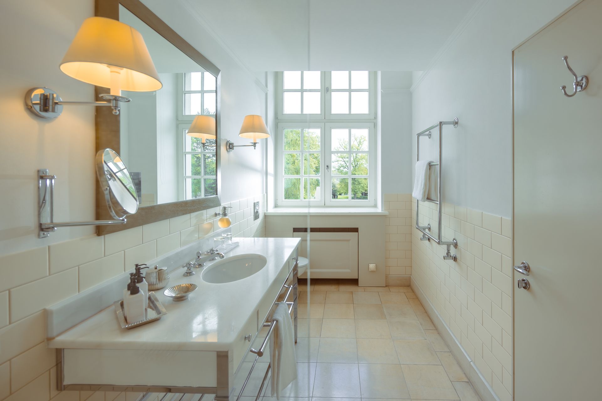 A bright, white bathroom with a vanity unit, a large mirror, wall sconces, and a window overlooking the garden.