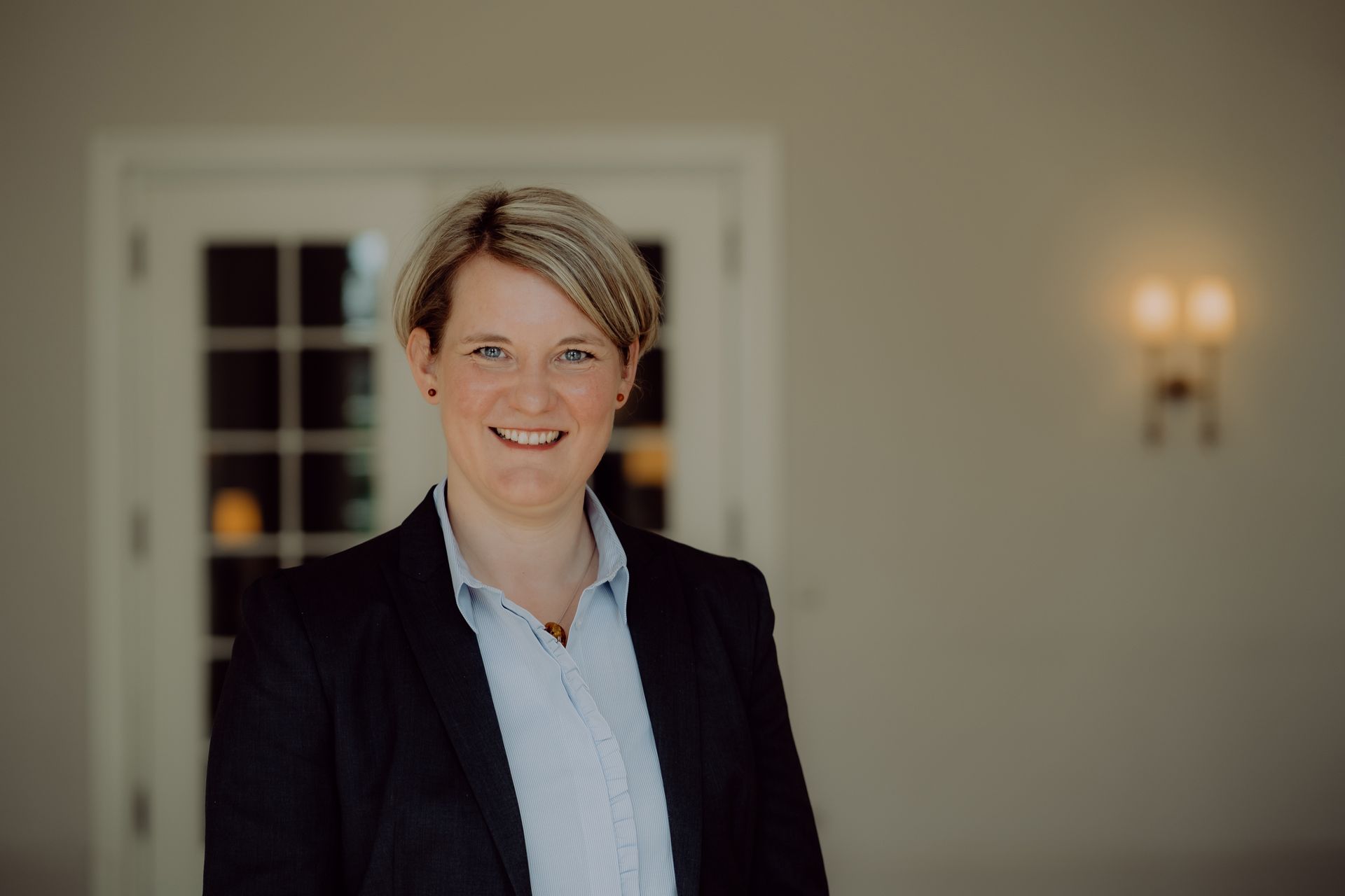 Frau im Blazer lächelt, steht in der Nähe der Flügeltüren.