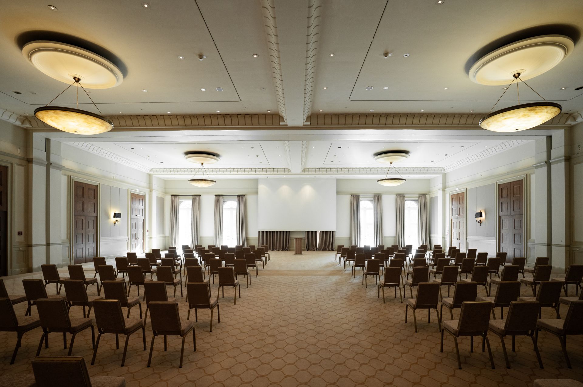 Großer, leerer Ballsaal mit Stuhlreihen vor einer Bühne. Beiger Teppichboden, dekorative Decke und Fenster.