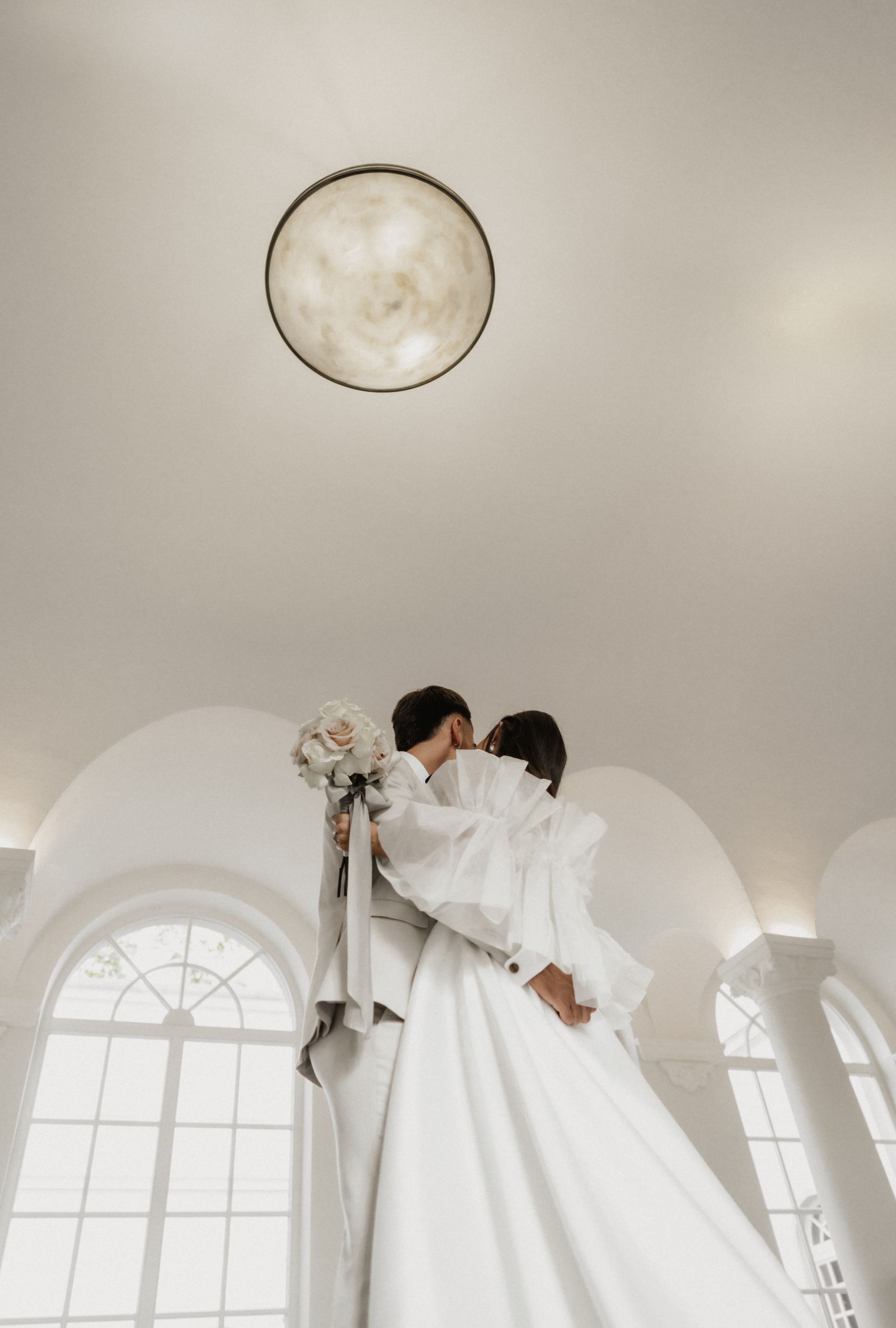 A couple is kissing in a white room with large windows, holding a bouquet of flowers.