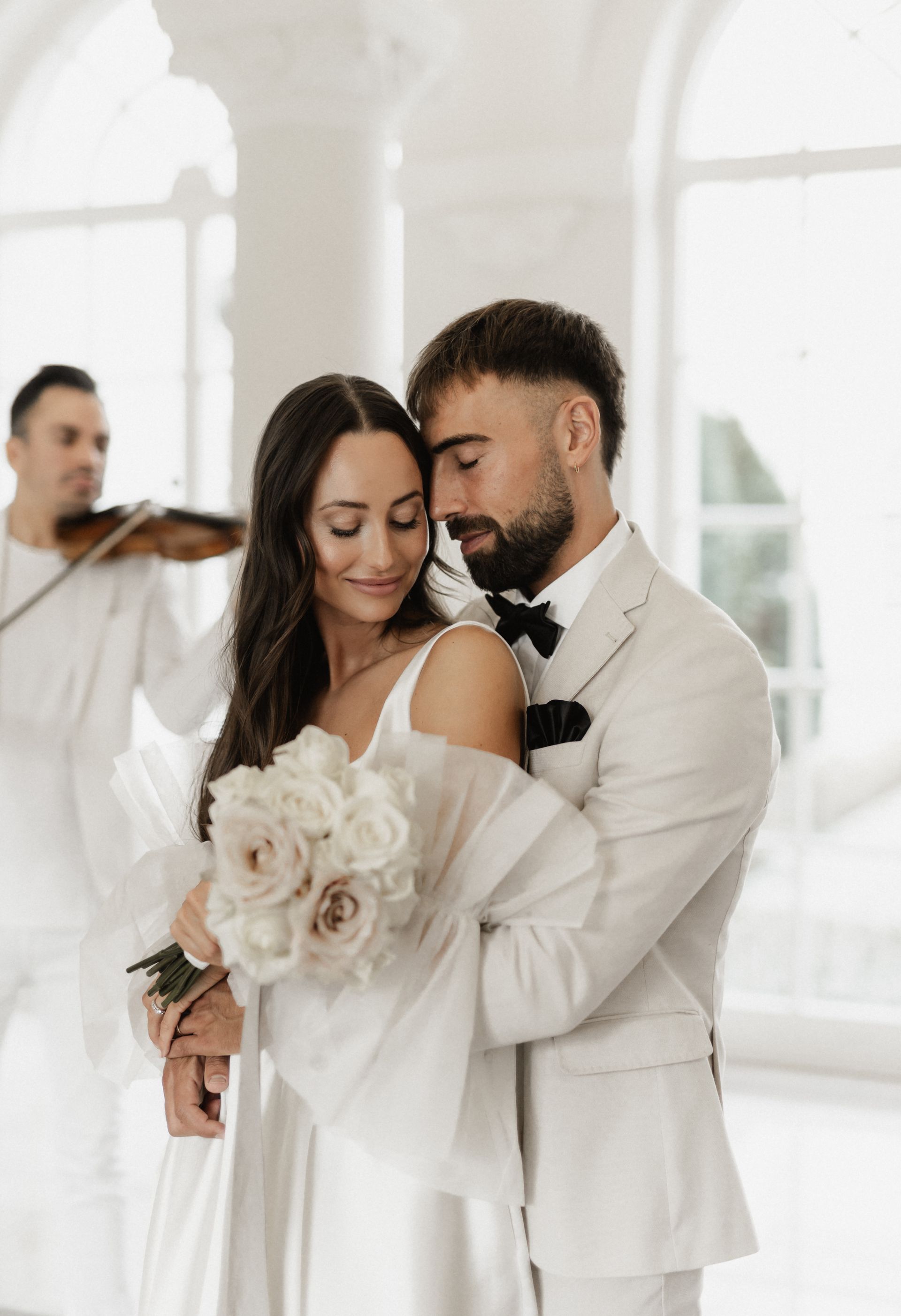 A bride and groom are embracing and holding flowers. A violinist is playing; the interior is white, and the lighting is soft.