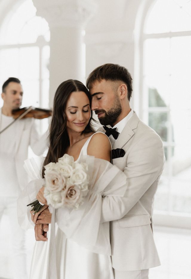 Ein Brautpaar umarmt sich und hält Blumen. Es spielt ein Violinist; der Innenraum ist weiß, die Beleuchtung sanft.