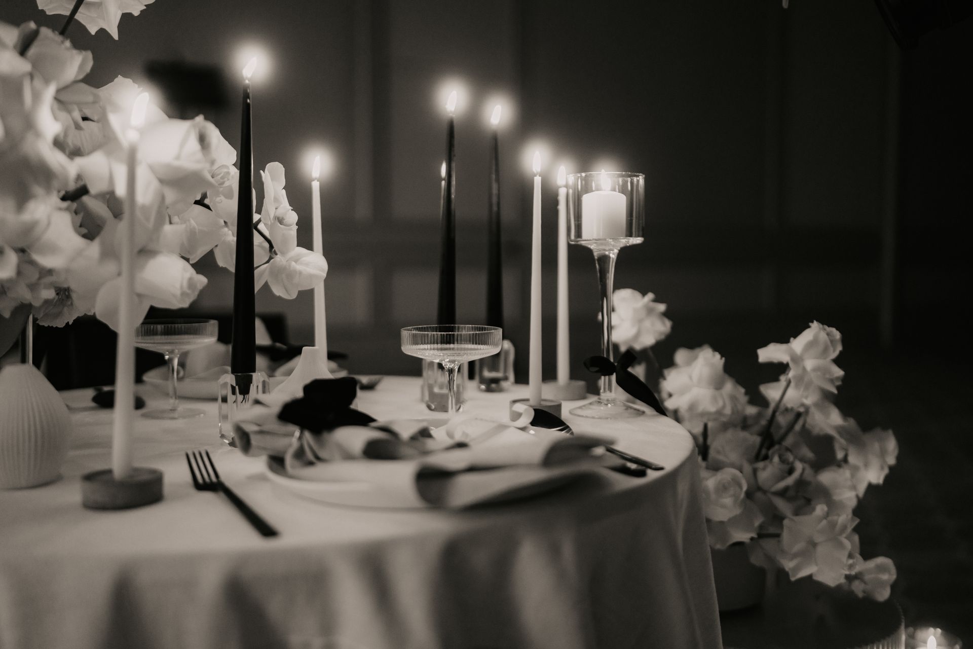Elegant gedeckter Tisch mit brennenden Kerzen und weißen Blumen, der eine romantische Atmosphäre in Schwarz-Weiß schafft.
