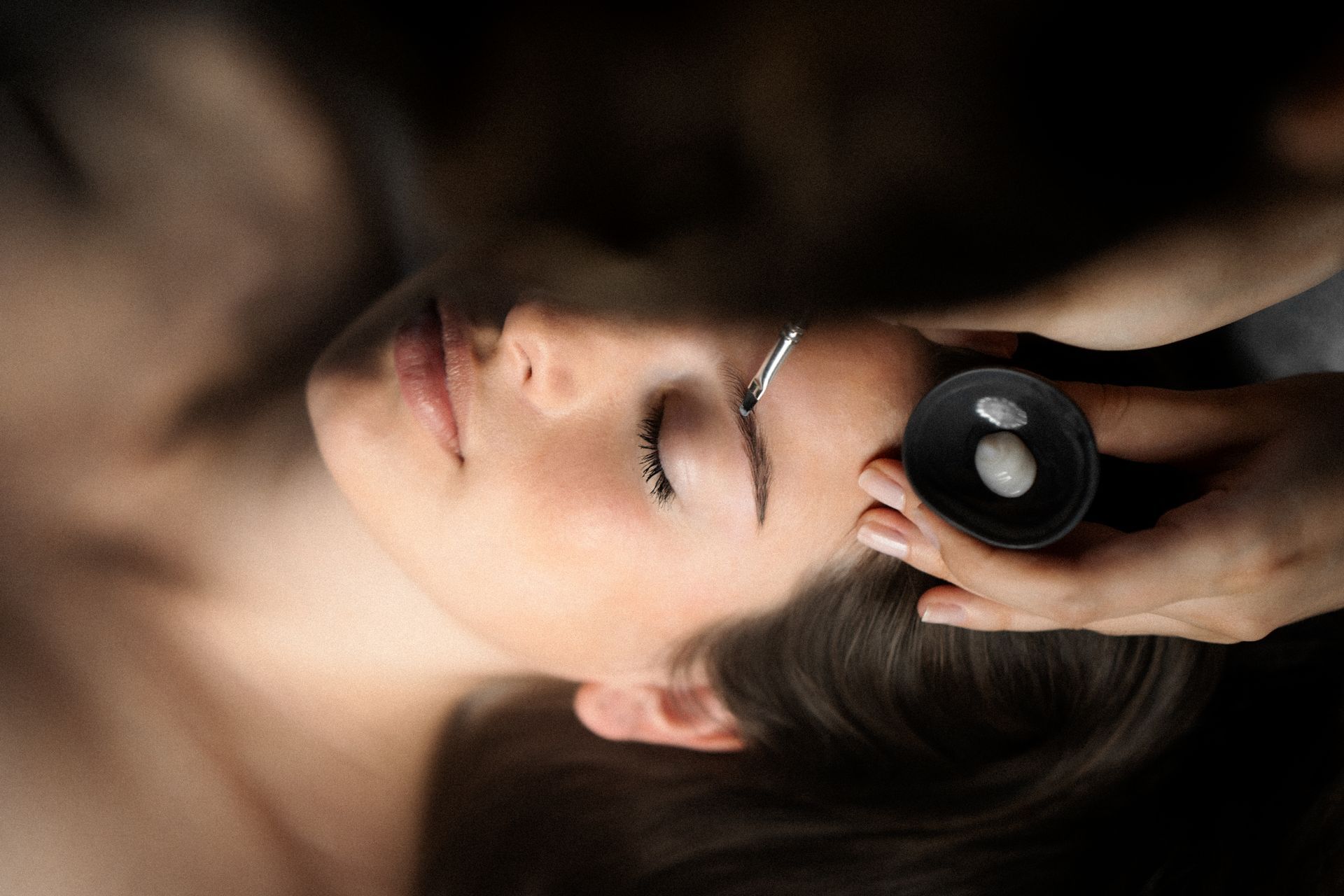 A woman undergoing a facial treatment, holding a container of white substance near her ear.