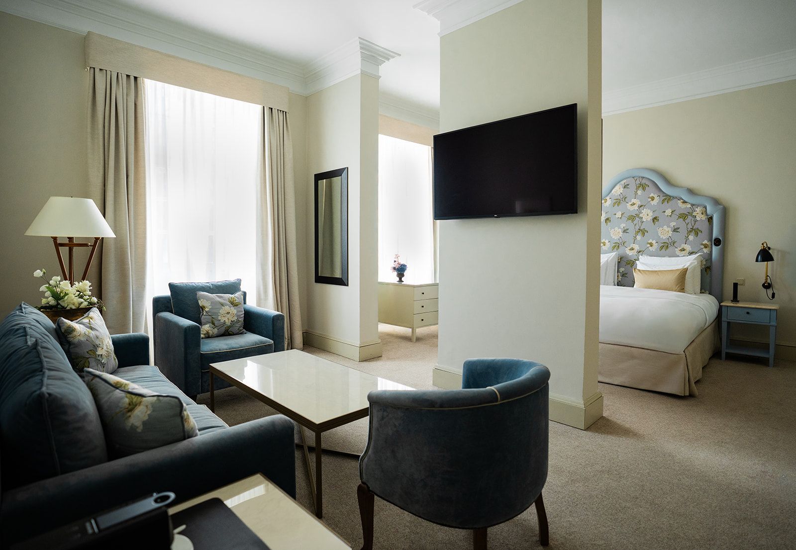 Hotel suite with a blue sofa, armchair, bed, and TV. Neutral colors, elegant design.