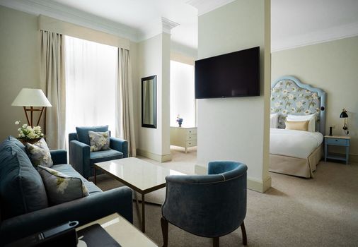 Hotelsuite mit blauem Sofa, Sessel und floralem Kopfteil. Beige Wände und Teppichboden.