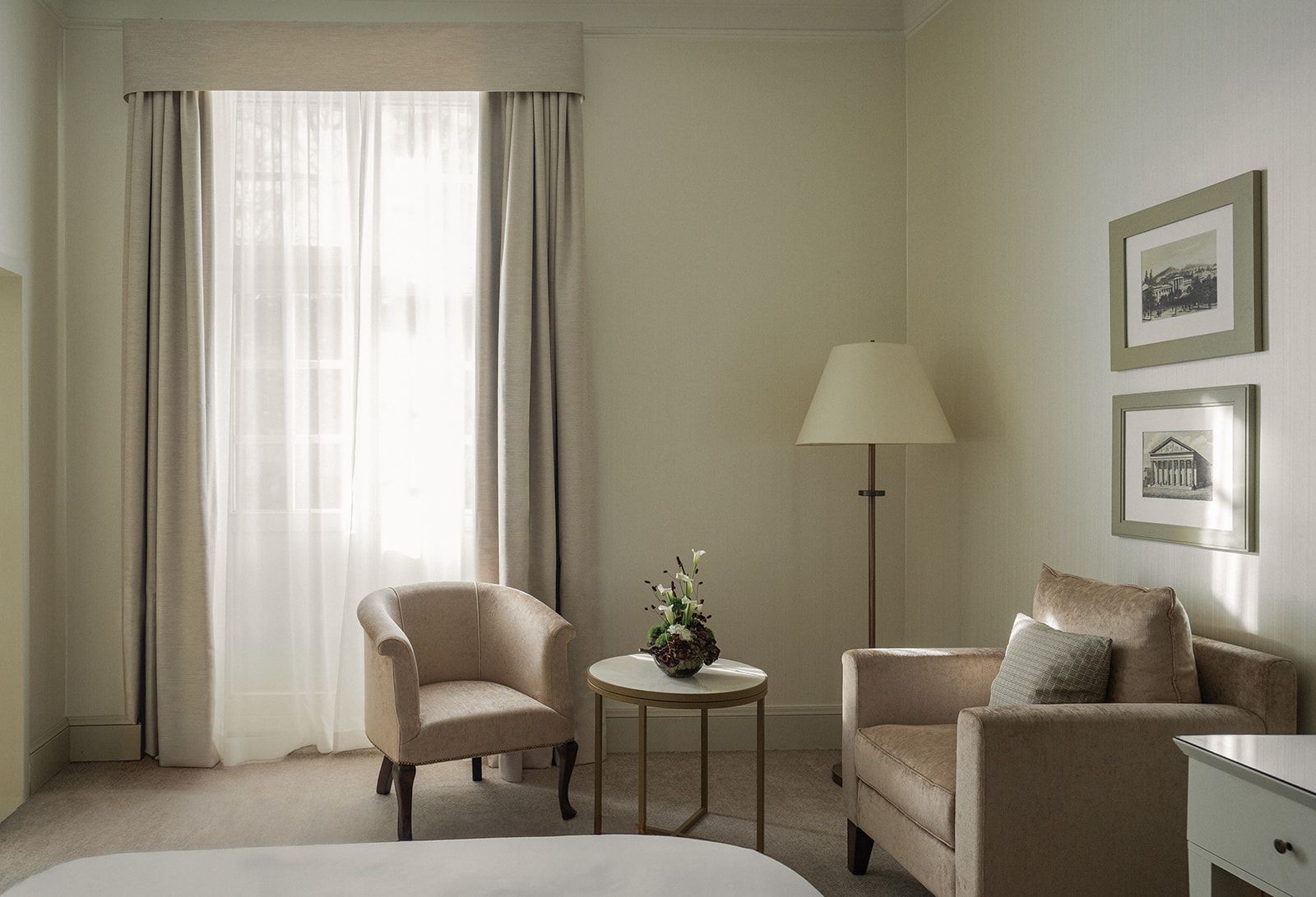 A cozy beige hotel room with a window, two armchairs, a side table with flowers, and framed pictures.