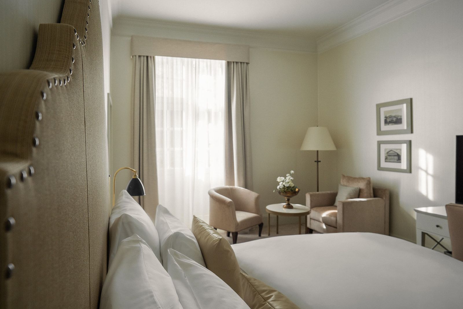A hotel bed with a high headboard, two armchairs around a round table, a floor lamp, and framed pictures on the wall.