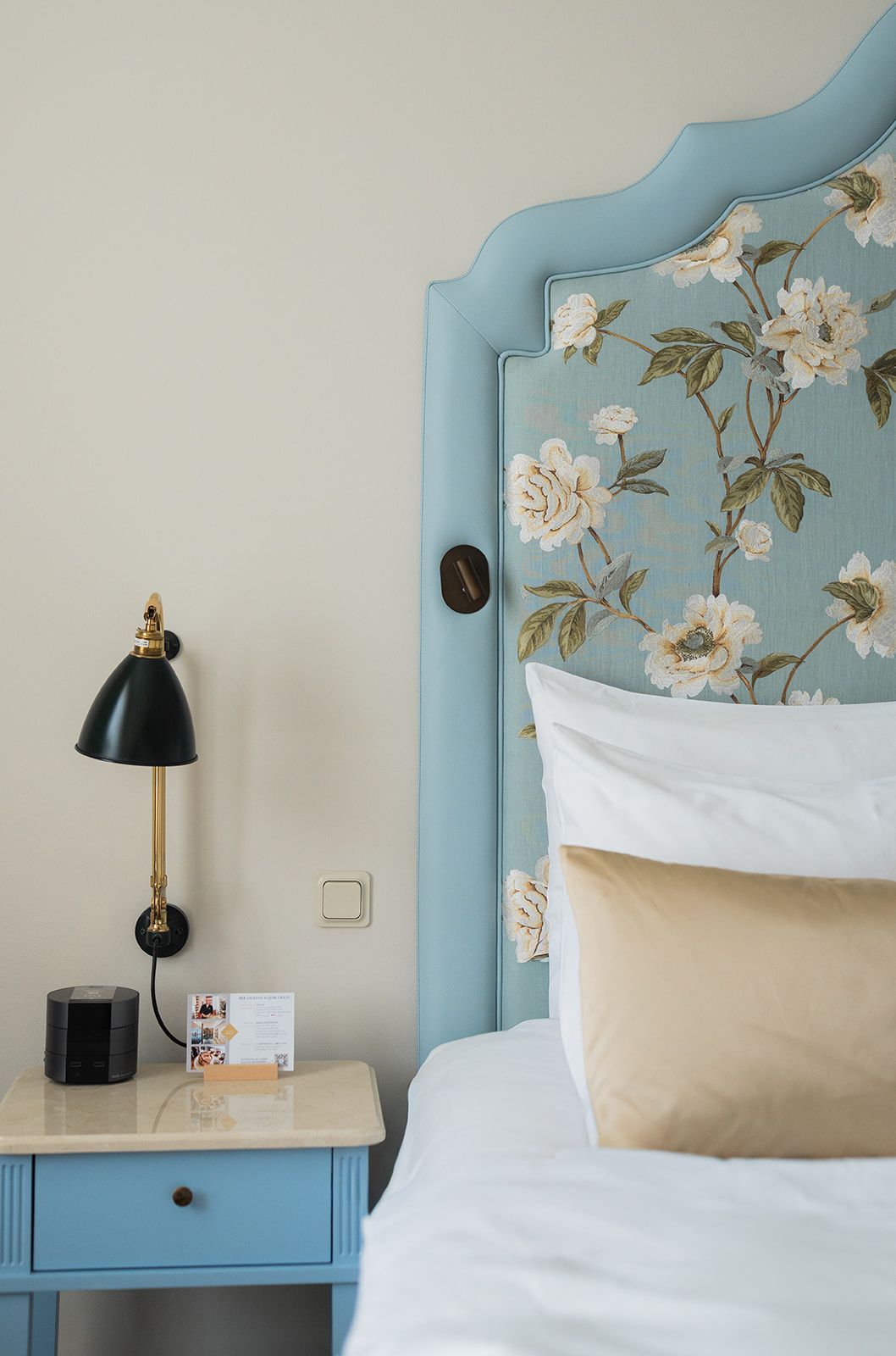 Schlafzimmerecke: blaues geblümtes Kopfteil, weiße Bettwäsche, goldenes Kissen, schwarze Lampe und blauer Nachttisch.