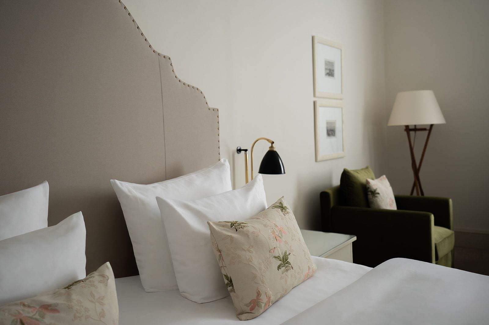 A hotel room featuring a bed with a light brown upholstered headboard, white pillows, patterned throw pillows, and a green armchair.