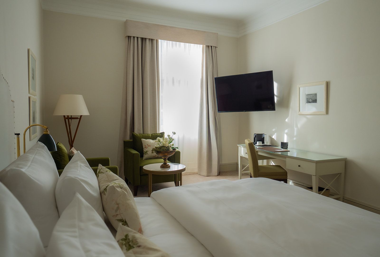 Schlafzimmer mit Bett, Schreibtisch, Sessel und Wandfernseher. Beige Wände, helle Vorhänge und eine Lampe.