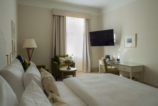 Schlafzimmer mit Bett, Schreibtisch, Sessel und Wandfernseher. Beige Wände, helle Vorhänge und eine Lampe.