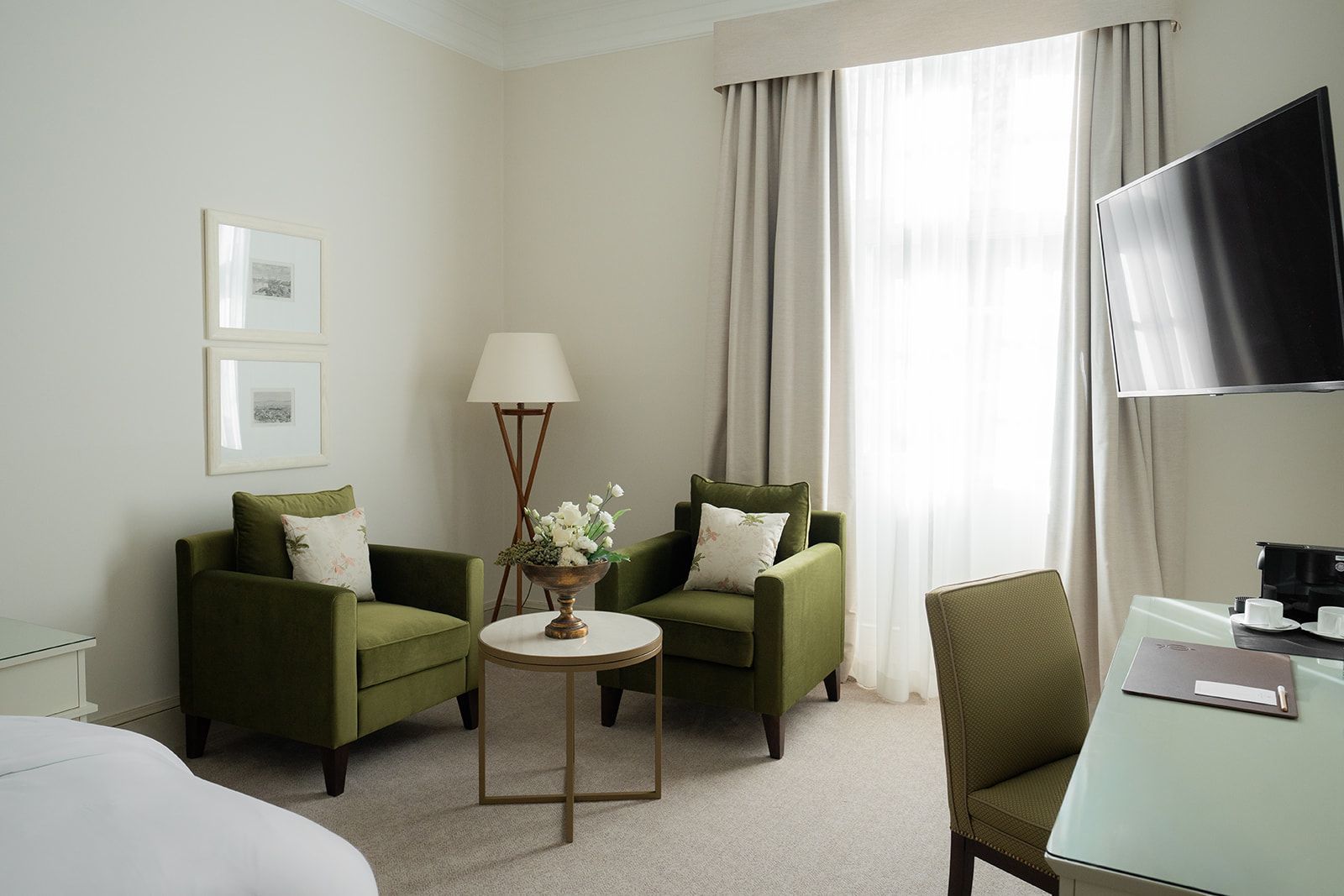 A hotel room with two green armchairs, a side table, a floor lamp, a desk, and a TV on the wall.