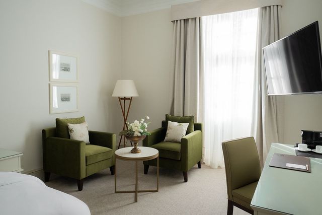 Hotelzimmer mit zwei grünen Sesseln, einem runden Tisch, Stehlampe, Schreibtisch und ein an der Wand montierter Fernseher.
