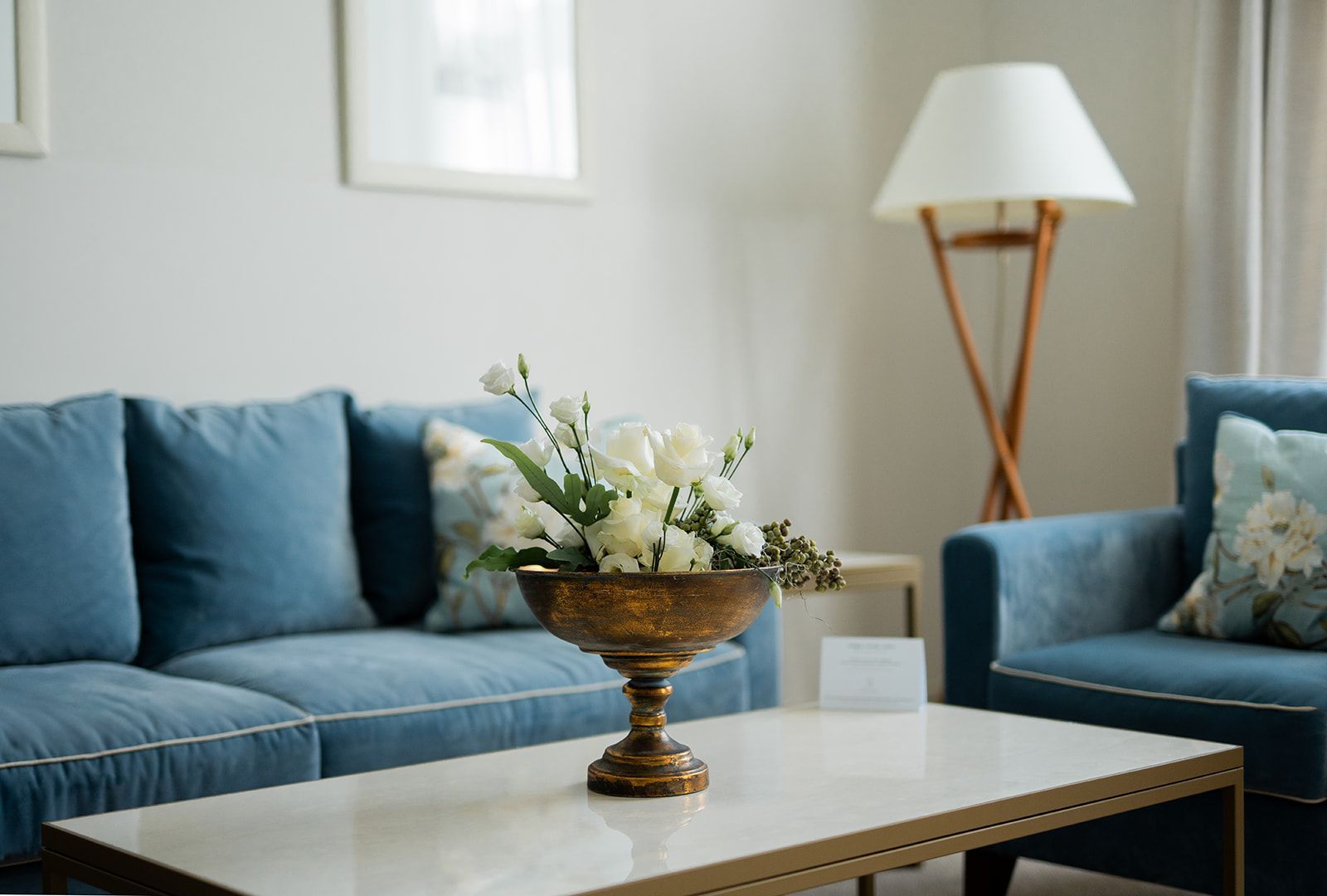 Blaues Sofa und Sessel mit weißen Blumen auf einem Couchtisch in einem lichtdurchfluteten Wohnzimmer.