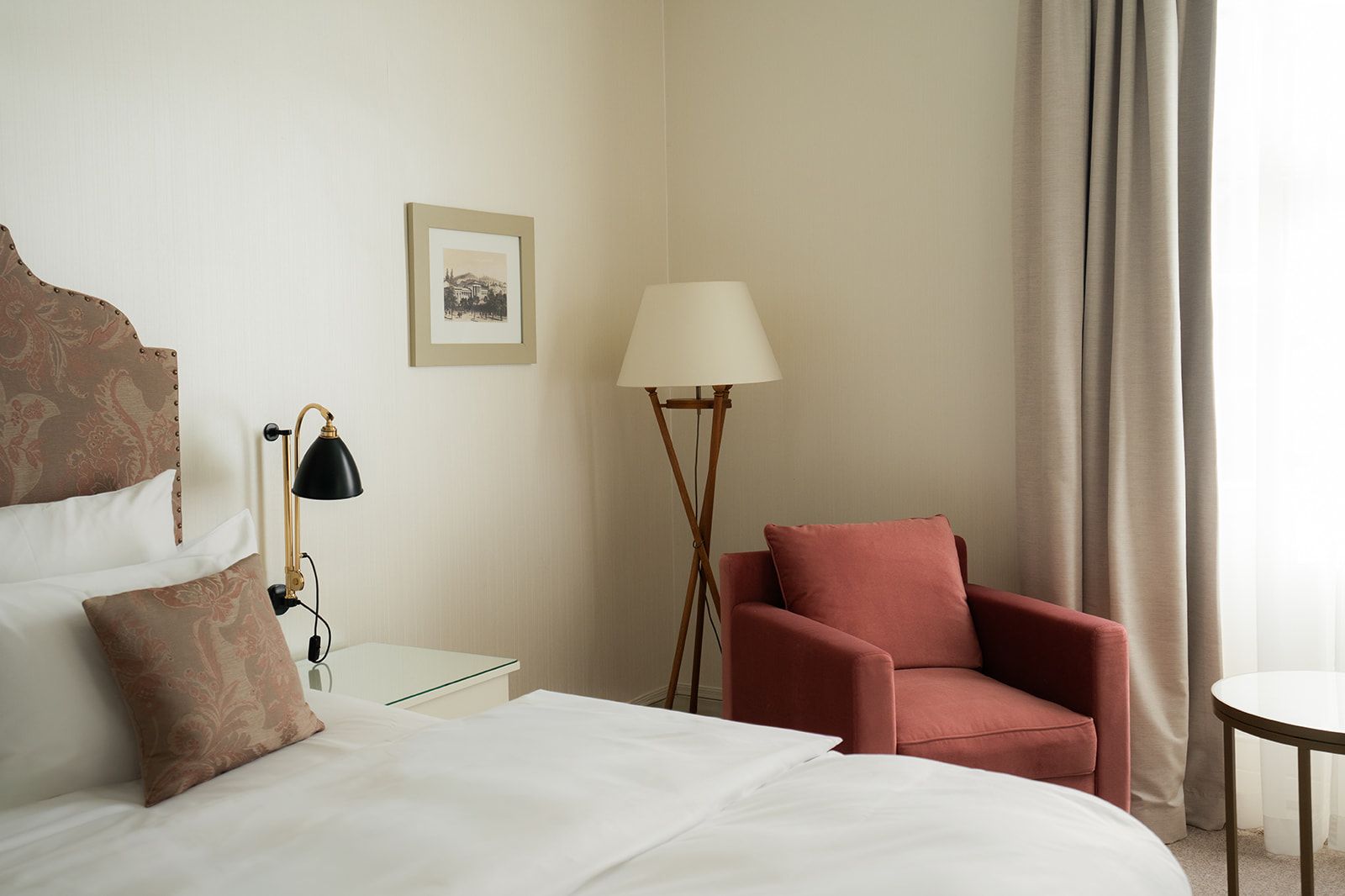 Ein Hotelzimmer mit Bett, braunem Zierkissen, Leselampe, Stehlampe, korallenfarbenem Sessel und beigefarbenen Vorhängen.