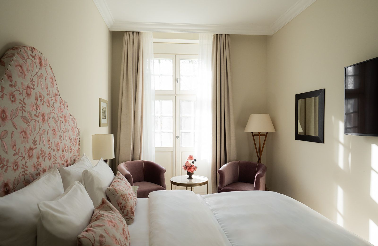 Schlafzimmer mit floralem Kopfteil, zwei Sesseln, einem Tisch und einem Fenster mit Vorhängen.