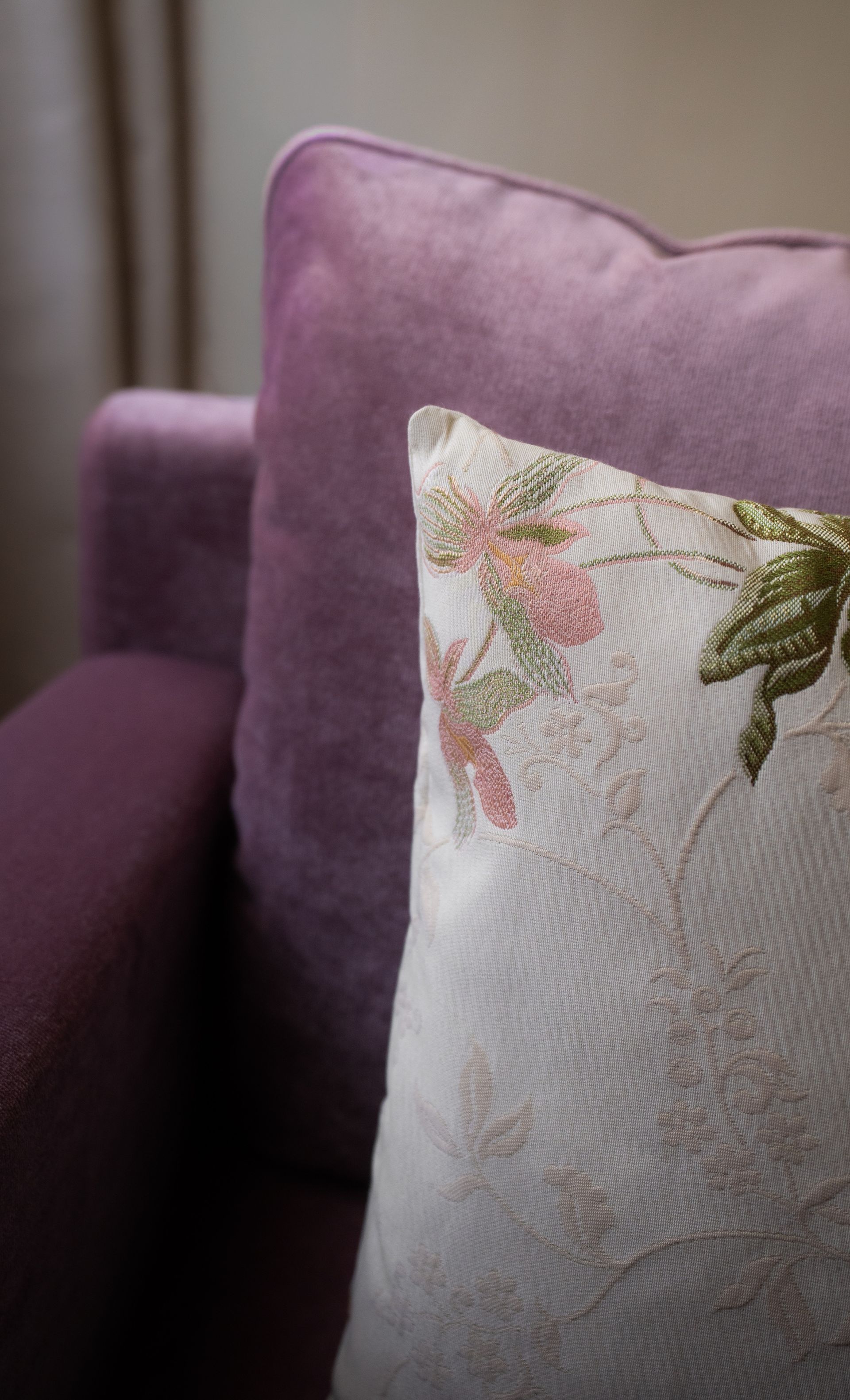 Ein strukturiertes, helles Kissen mit Blumenstickerei lehnt an einem plüschigen, tiefvioletten Sessel.