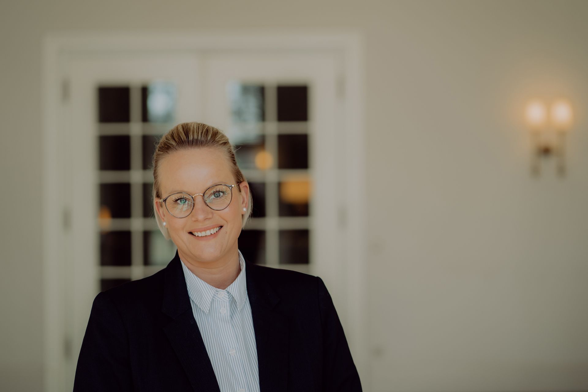 Frau mit Brille lächelt, sie trägt einen schwarzen Blazer. Innenraum mit weißen Türen, Wandleuchte und hellen Wänden.