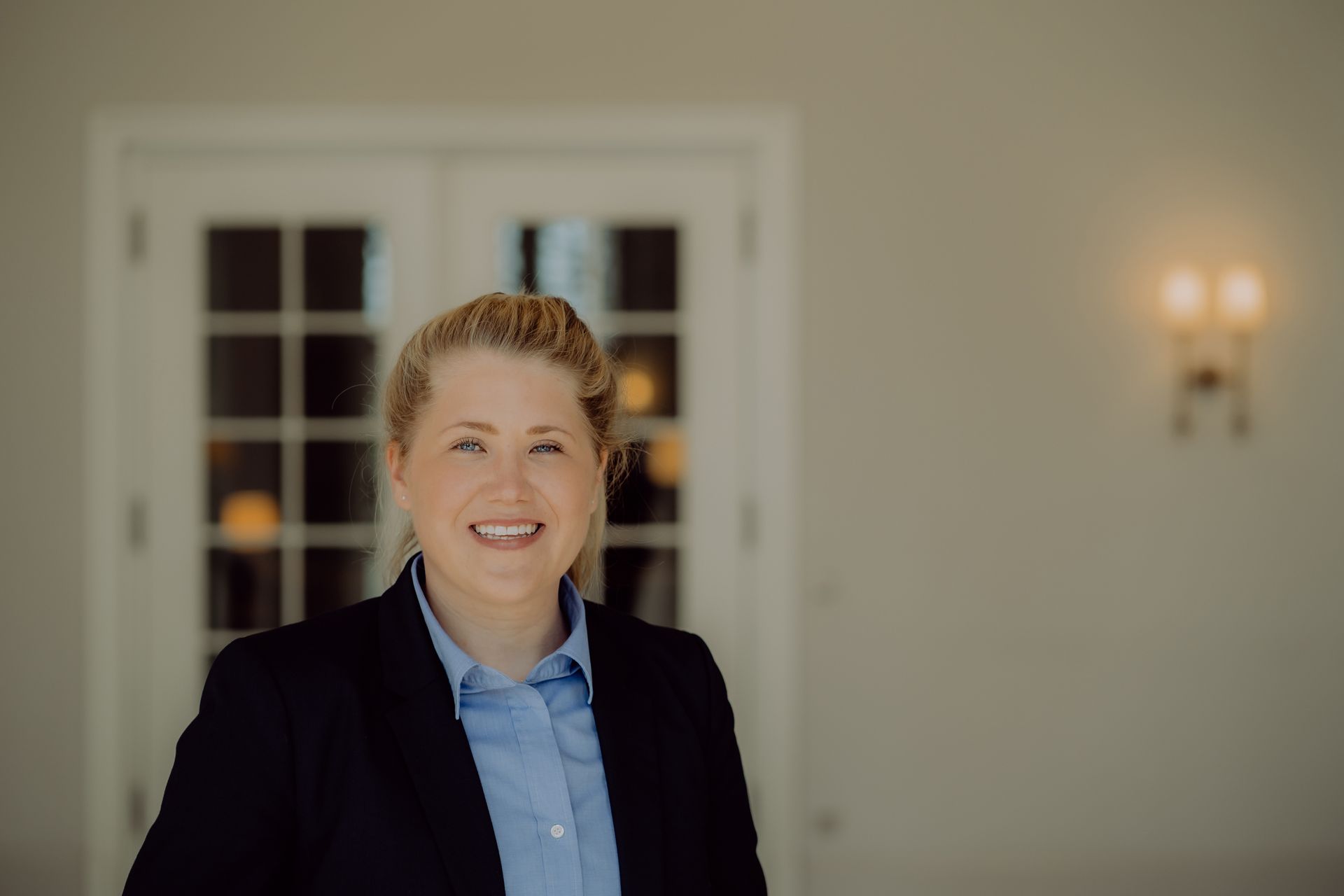 Eine Frau im Blazer lächelt vor weißen Flügeltüren.