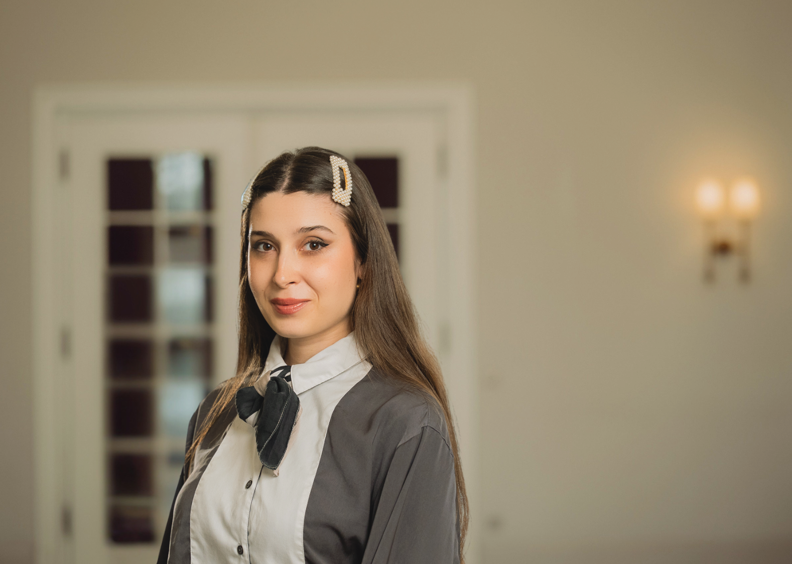 Frau mit langen Haaren in Schuluniform, die in einem Innenraum in der Nähe einer Tür und einer Wandleuchte steht.