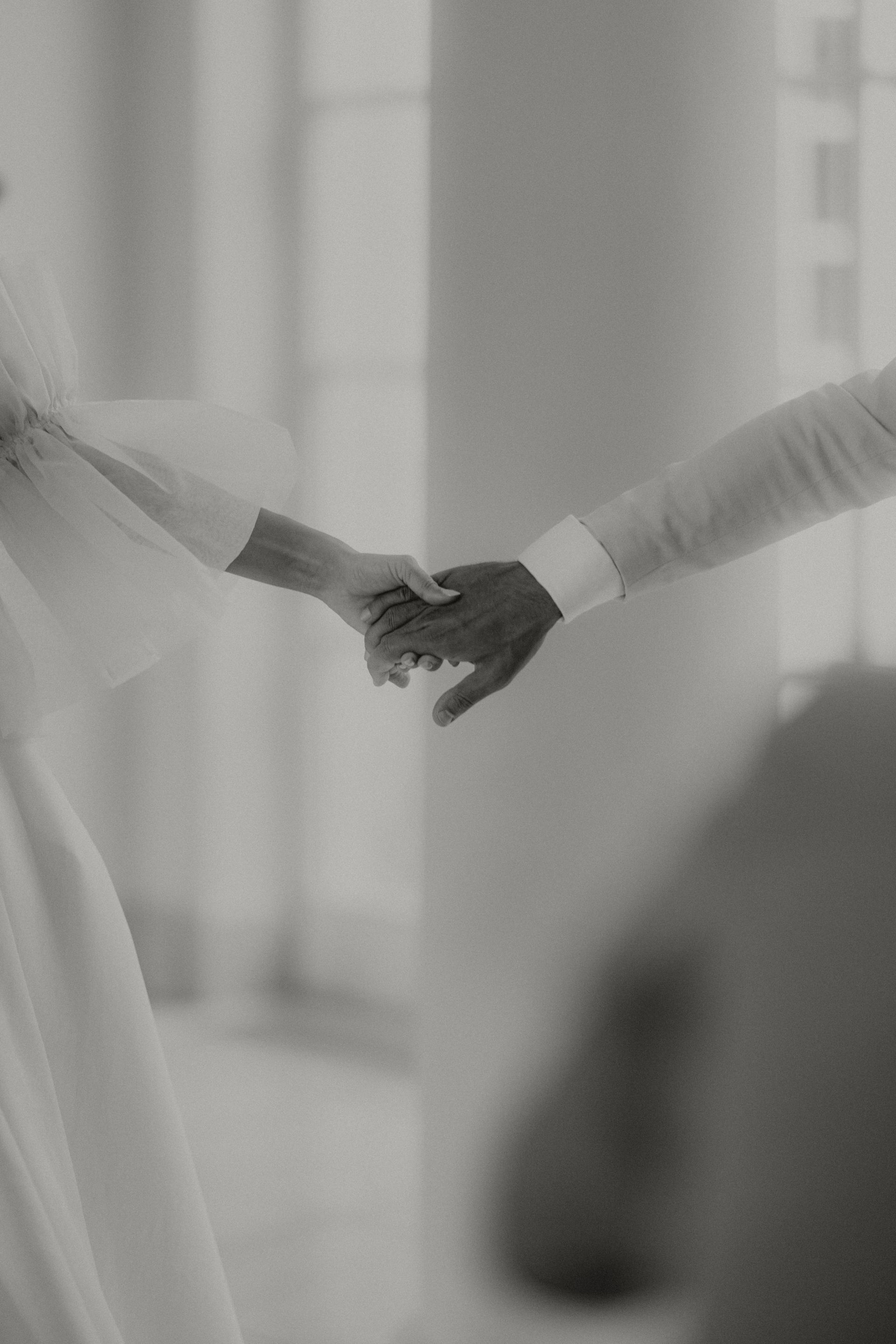Zwei ineinander verschränkte Hände, möglicherweise während einer Zeremonie. Weicher Fokus bei neutraler Beleuchtung.