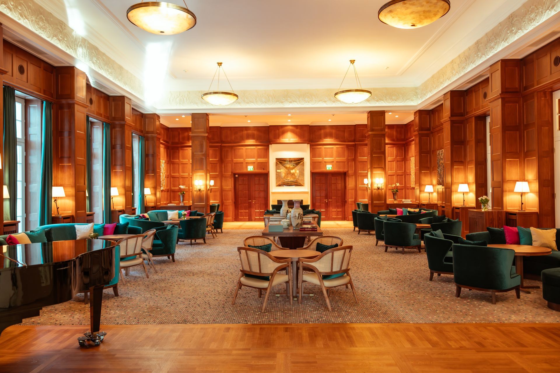 Eine elegante Hotellounge mit holzgetäfelten Wänden, türkisfarbenen Samtsesseln, runden Tischen und warmem Deckenlicht.