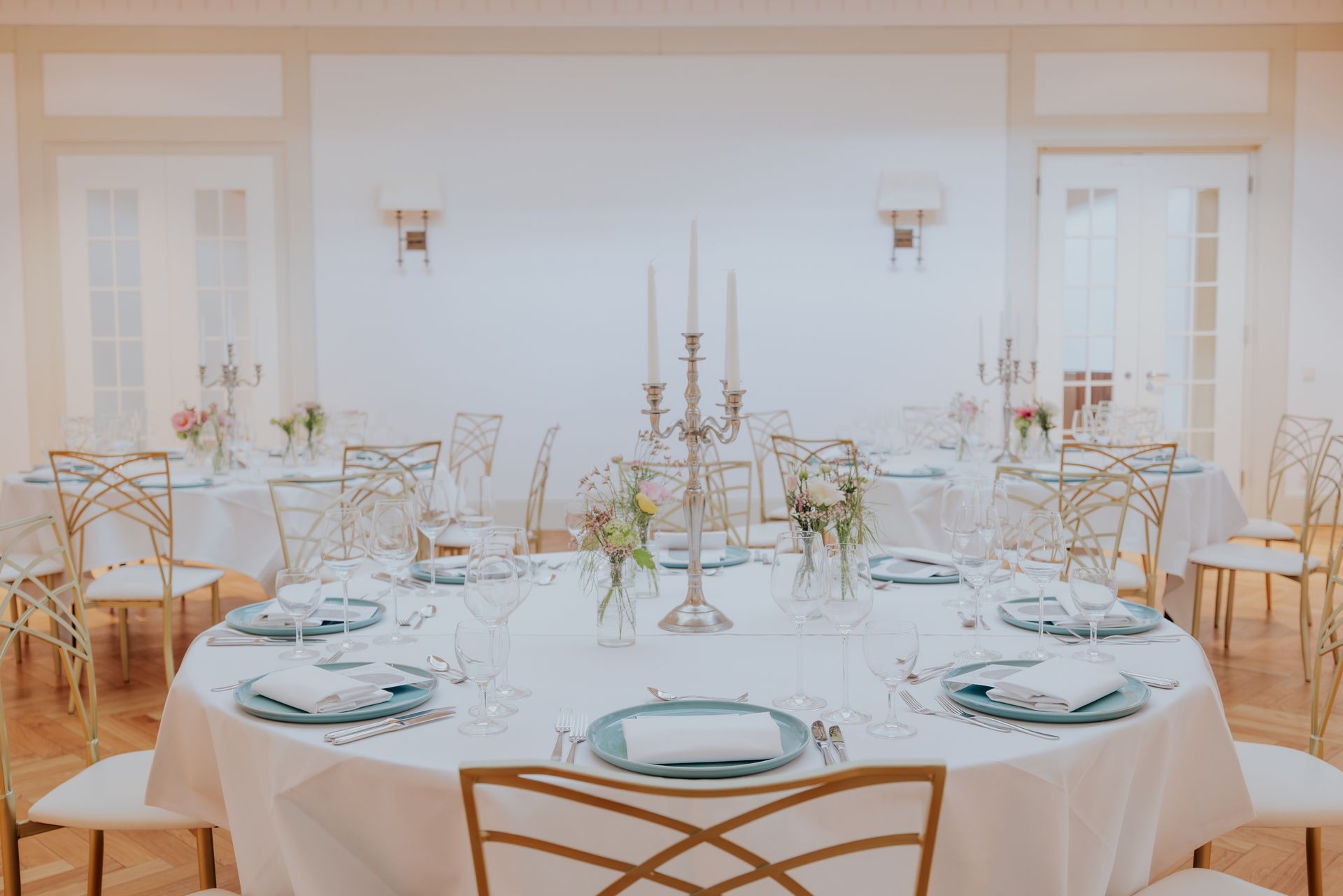 Ein formelles Esszimmer mit festlich gedeckten Tischen; weiße Tischdecken, goldene Stühle und florale Gestecke.