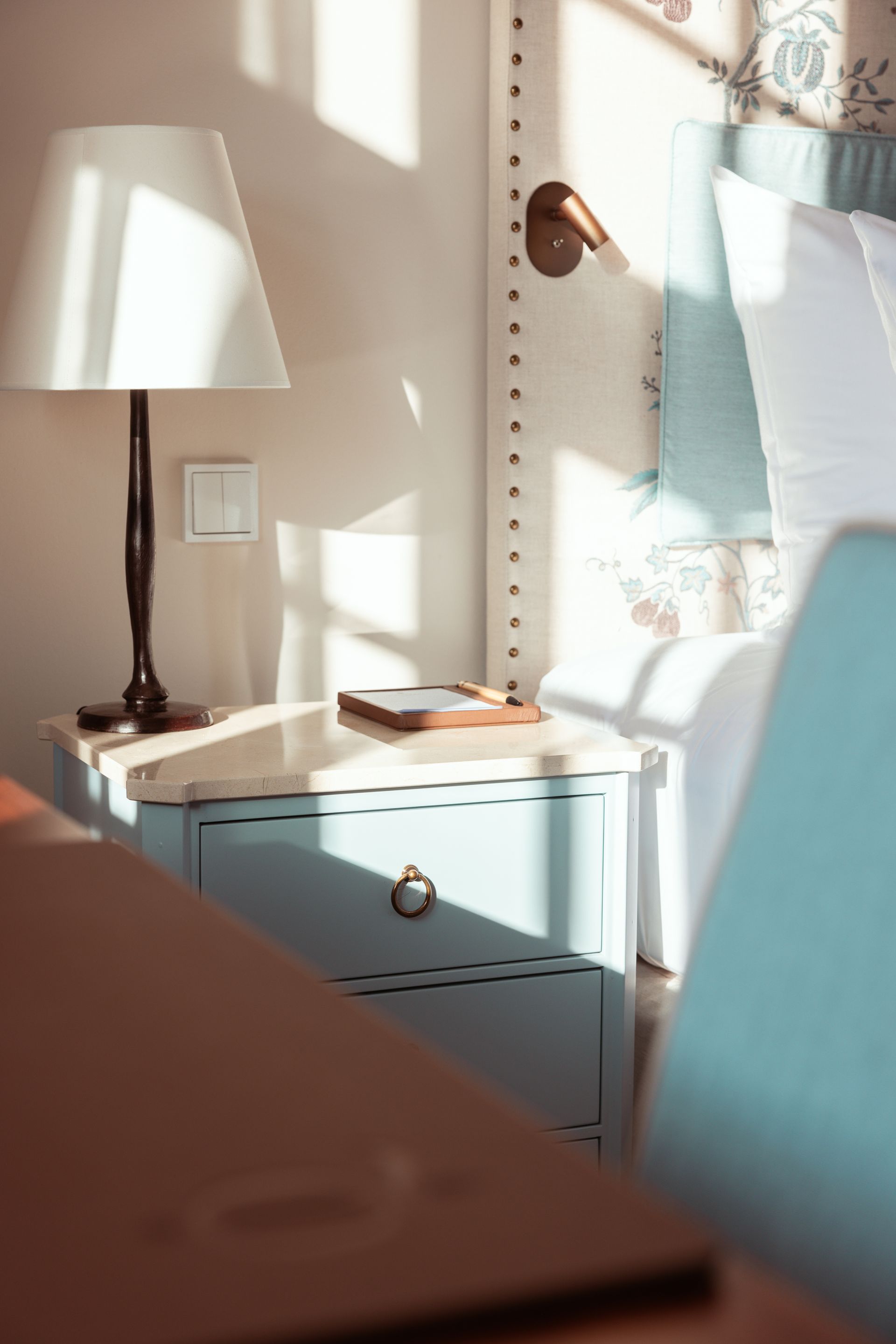 A light blue nightstand with a lamp and a book next to the bed in a sun-drenched, elegantly furnished bedroom.
