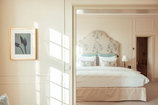 Schlafzimmerinterieur mit floralem Kopfteil, gerahmten Bildern und hellem Sonnenlicht.