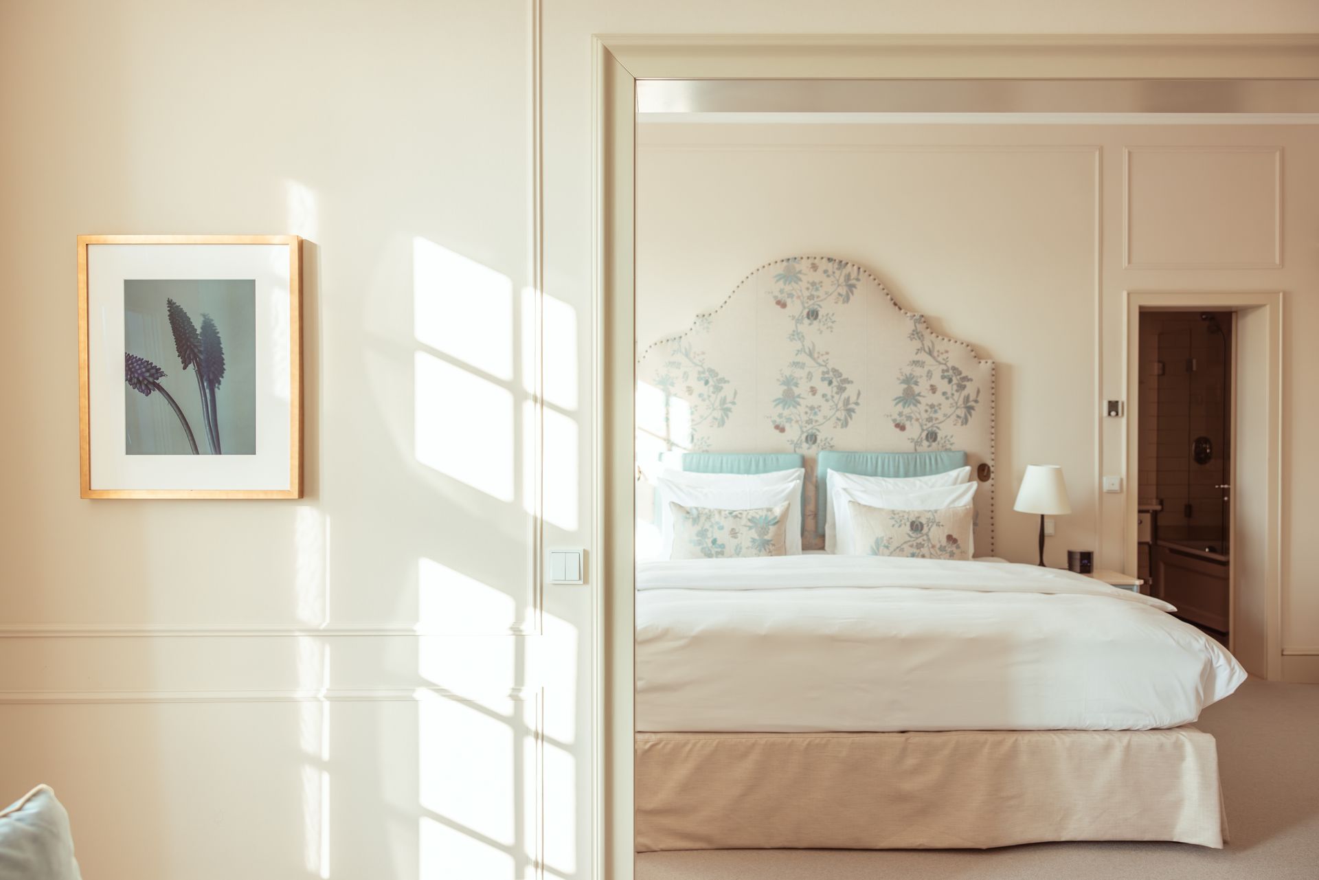 Schlafzimmerinterieur, florales Kopfteil, gerahmte Bilder, Sonnenlicht durchs Fenster.