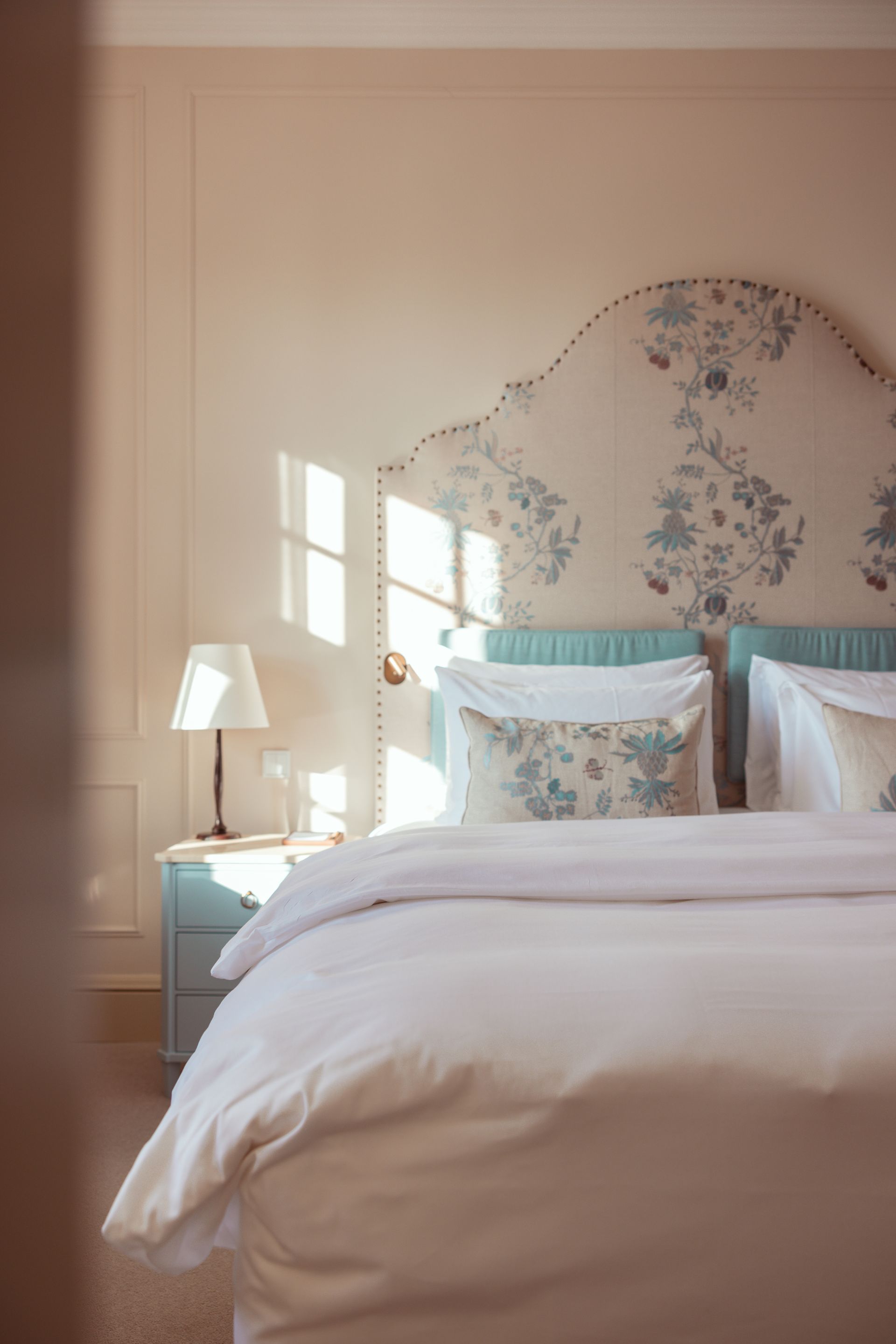 Schlafzimmer mit einem großen Bett, einem floralen Kopfteil, blauen Akzenten und Sonnenlicht, das durch ein Fenster strömt.