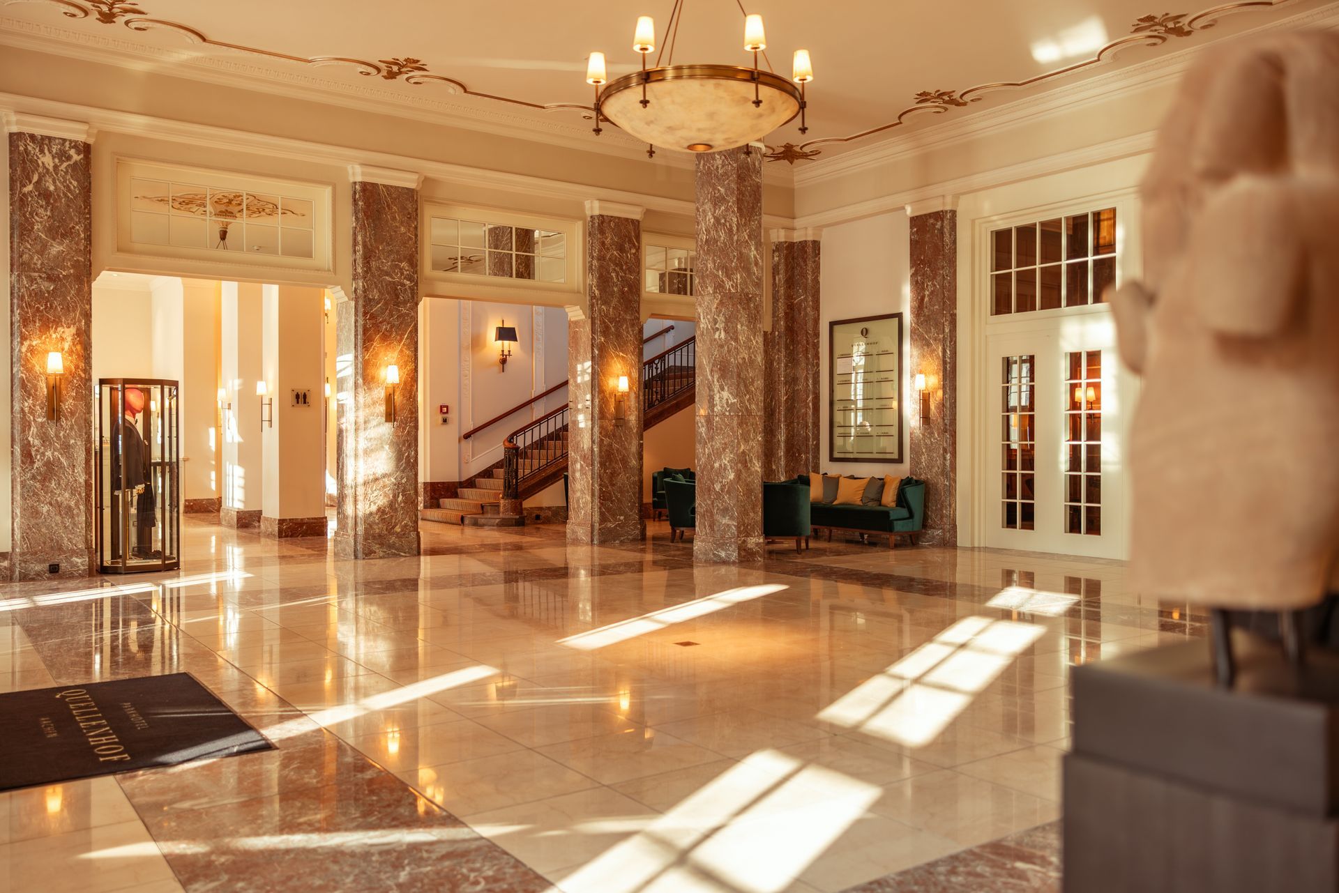Eine elegante Hotellobby mit Marmorböden, Ziersäulen, einer prunkvollen Treppe, einem großen Kronleuchter und einer Skulptur.