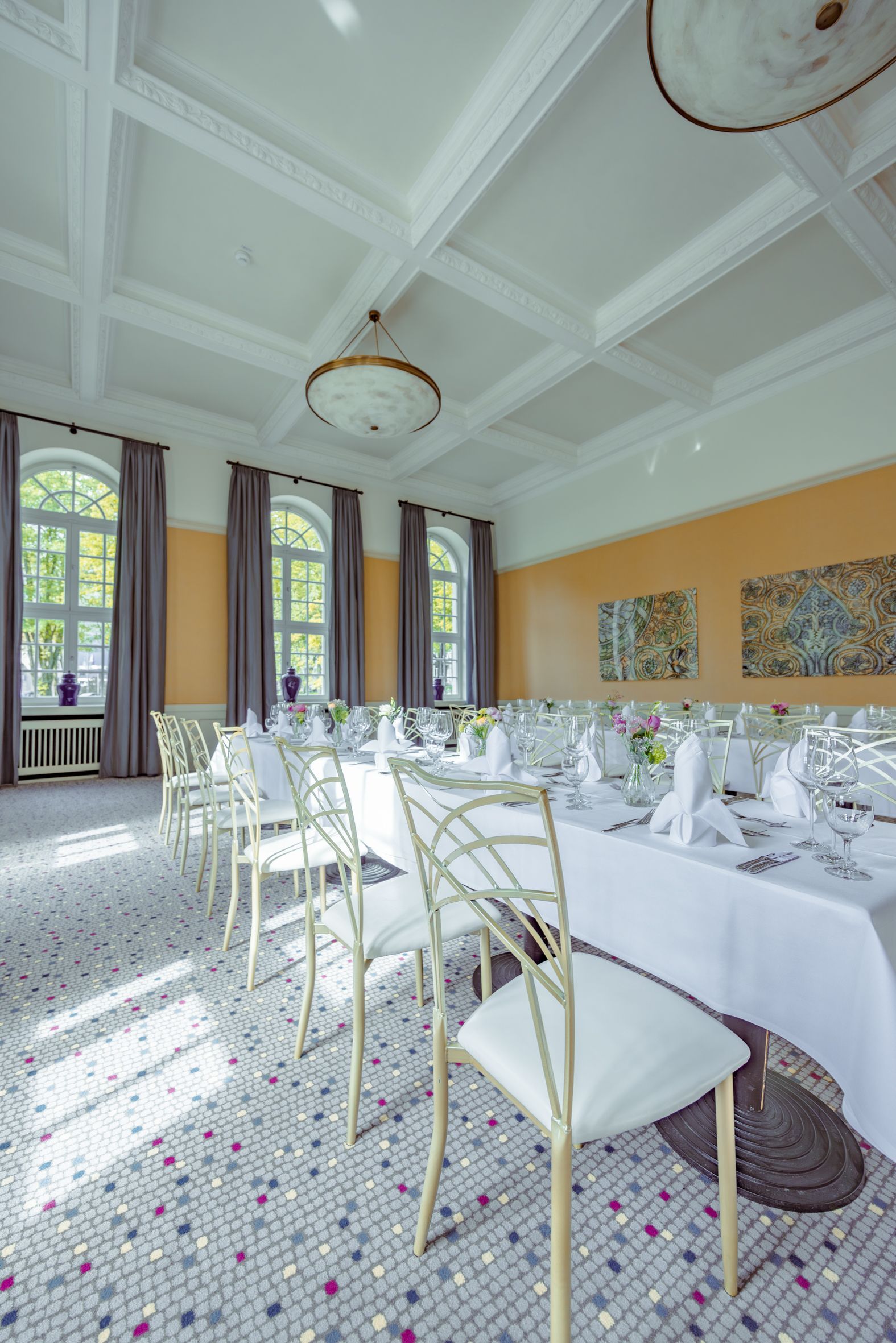A dining room set for a banquet. White chairs and tables on a patterned carpet, arched windows, and a decorative ceiling.
